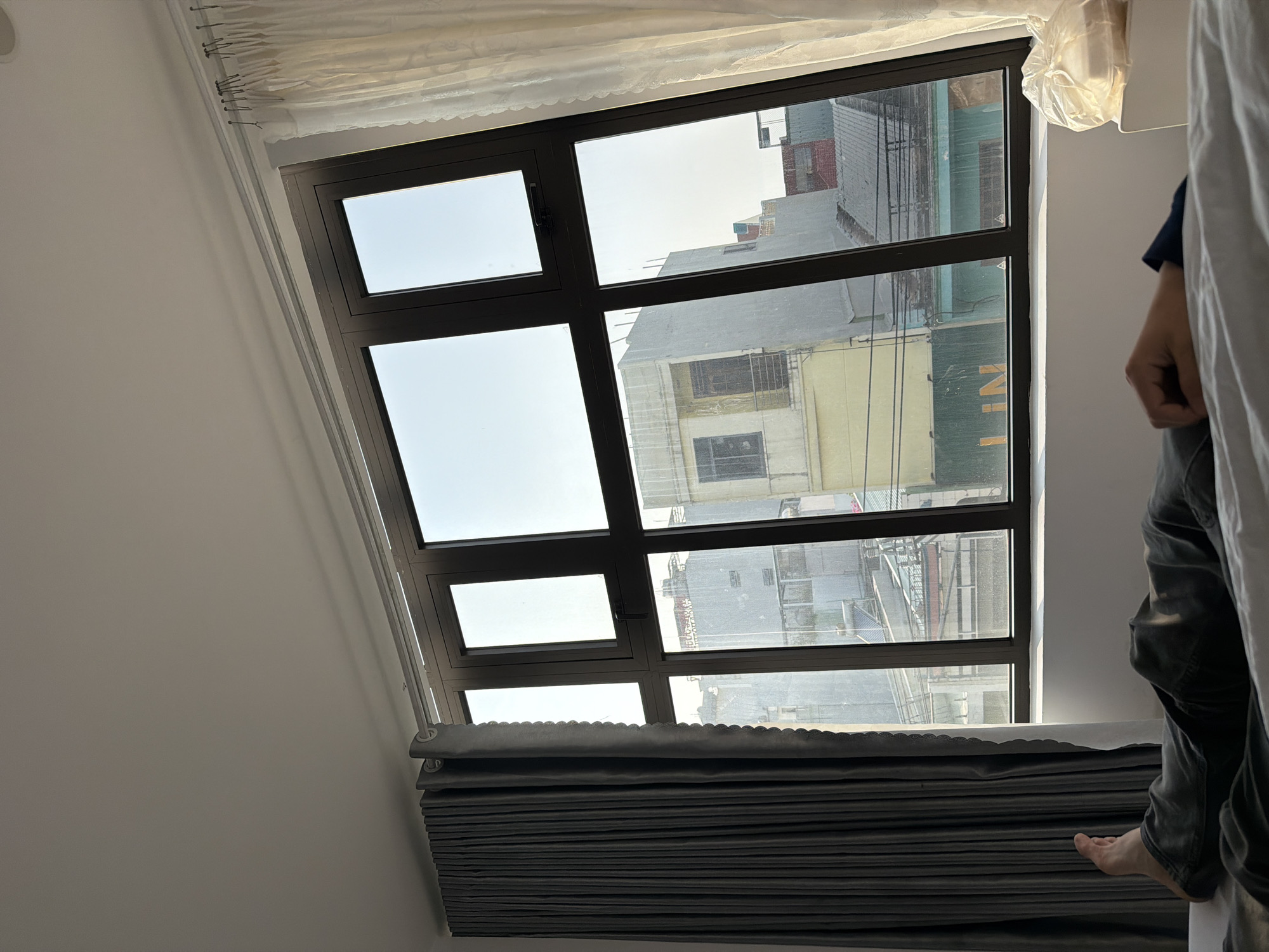 A skewed, interior shot shows a tilted, multi-paned window overlooking a city, possibly in Quy Nhon Ward, Vietnam. The view through the window includes buildings with varied architecture, from taller structures to lower-rise residential or commercial buildings. The sky is visible through the window panes, suggesting daylight conditions.
A person's partial presence is visible on the right side of the frame. Only a person’s partially-visible foot, leg in gray pants, and arm with a blue shirt are visible behind a white curtain.
Gray, pleated curtains are rolled up at the bottom of the window. White, sheer curtains are at the top of the window. The window frame itself is dark, possibly black or dark brown. The wall adjacent to the window is a light color, possibly white or light gray. The photo is taken from a low angle, looking up at the window.