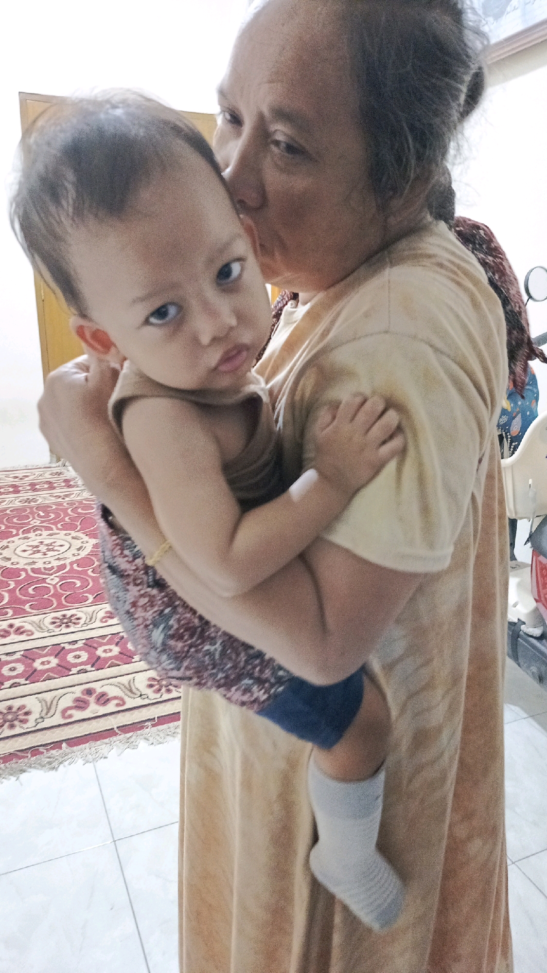 A middle-aged Indonesian woman, with dark hair pulled back in a bun and wearing a light brown dress, is holding a baby. She is kissing the baby on the forehead. The baby, with wide, curious eyes and light brown hair, is wearing a brown sleeveless shirt and blue shorts, with a colorful patterned fabric wrapped around its waist and legs. The baby's left foot is visible, wearing a white sock. They are standing on a tiled floor with a patterned rug to the left. In the background, a door and a portion of a white chair are visible. The lighting suggests an indoor setting.