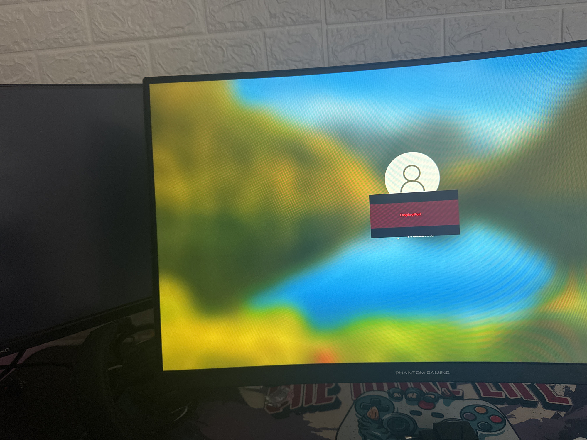 The image shows a computer setup. There are two monitors, one to the left which is mostly out of view, and a curved monitor in the center displaying the Windows login screen. A red error message overlay says "DisplayPort." The monitor's bottom bezel reads "PHANTOM GAMING." Below the monitor is a patterned desk mat featuring what appears to be a gaming controller design. The background includes a white brick wall. There is no information related to Saedinenie, Bulgaria.