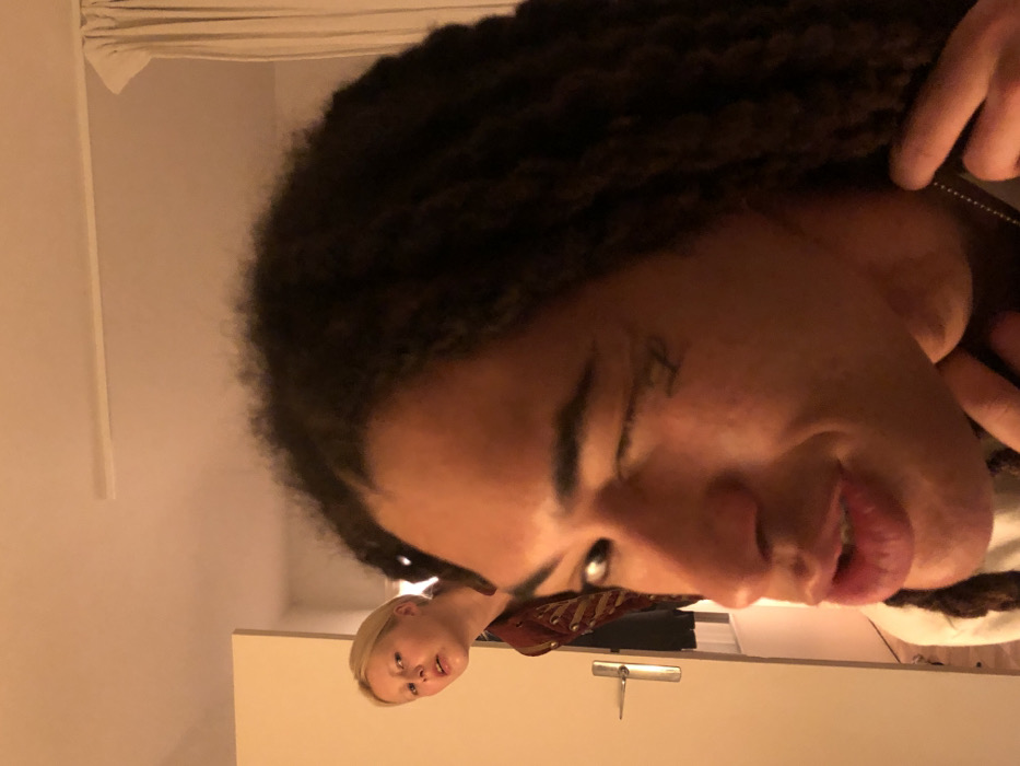 A selfie-style, tilted image shows a person with dark, curly hair and medium brown skin. They are smiling slightly with their mouth open. A second person with short, light blonde hair is also visible, looking up with a neutral expression. A white wall with a horizontal surface and silver hardware is in the background. A beige curtain hangs in the top left corner. The lighting is warm and soft.