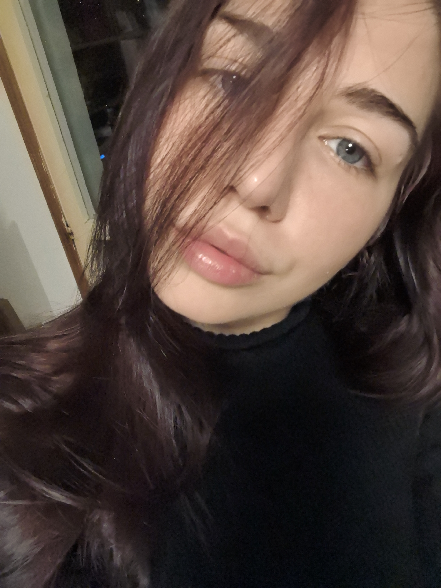A close-up, matter-of-fact portrait captures a single female individual with light skin, blue eyes, and long, dark brown hair with reddish undertones, a portion of which covers the left side of her face. Her lips appear full and glossy. She is wearing a black, ribbed turtleneck. The subject is indoors, facing the camera. A light-colored window frame is visible on the left side of the background. Beyond the window, the exterior is dark and blurred, consistent with night, with a single faint blue light source identifiable in the distance. The image contains no visual elements to specifically identify the location as Stara Zagora, Bulgaria.