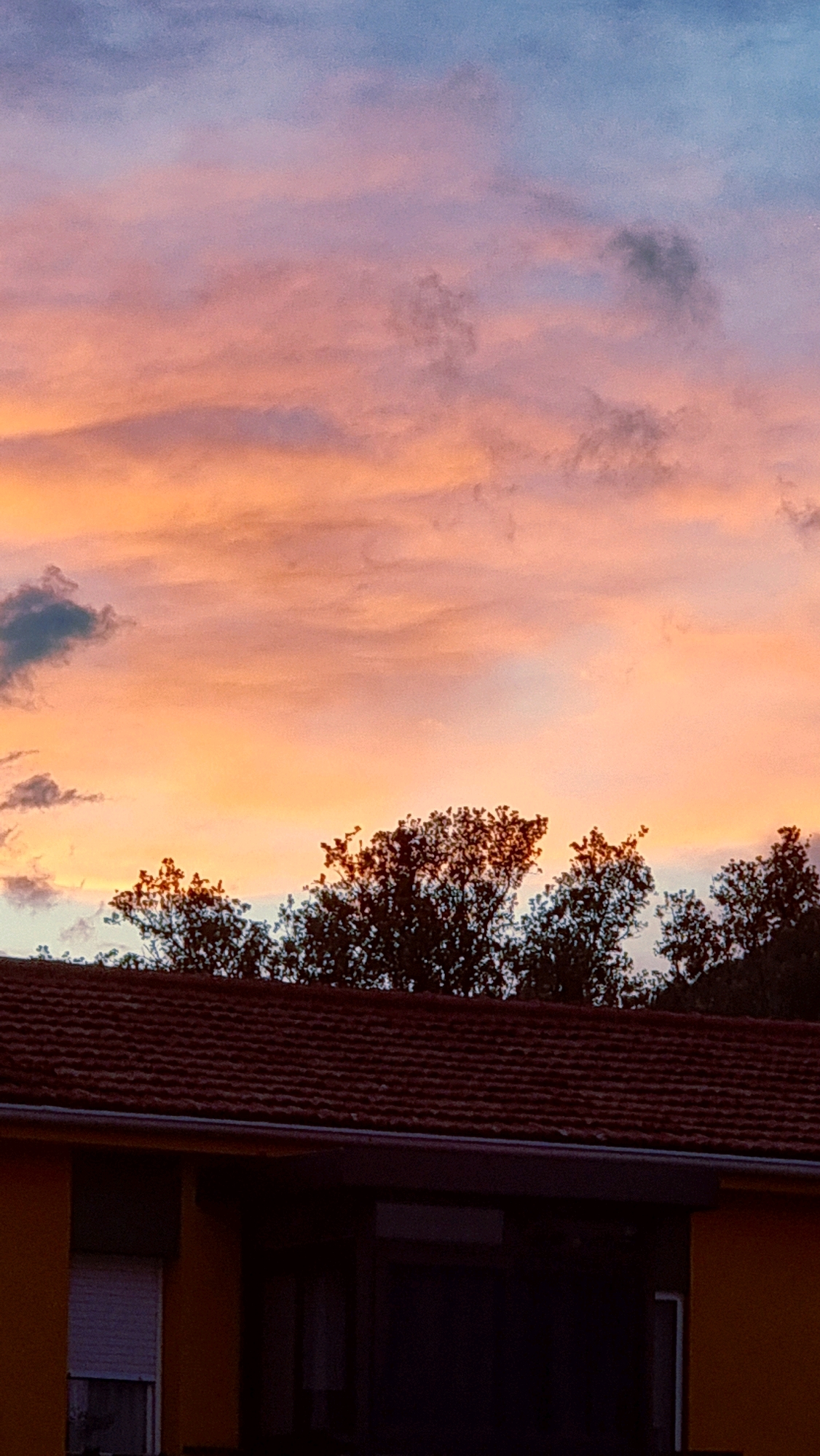 The image captures a sunset sky over the town of Sant Joan Les Fonts, Spain. The sky is filled with a gradient of colors, transitioning from soft pinks and oranges near the horizon to a light blue at the top. Scattered clouds, some appearing darker and more defined, are visible against the colorful sky. Silhouetted against the lower part of the sky are the tops of trees, their branches creating intricate patterns. In the foreground, the dark, tiled roof of a building is prominent, with a portion of a yellow wall visible on the left. A white-shuttered window can be seen on the yellow wall. The overall scene depicts a residential area under a dramatic twilight sky.
