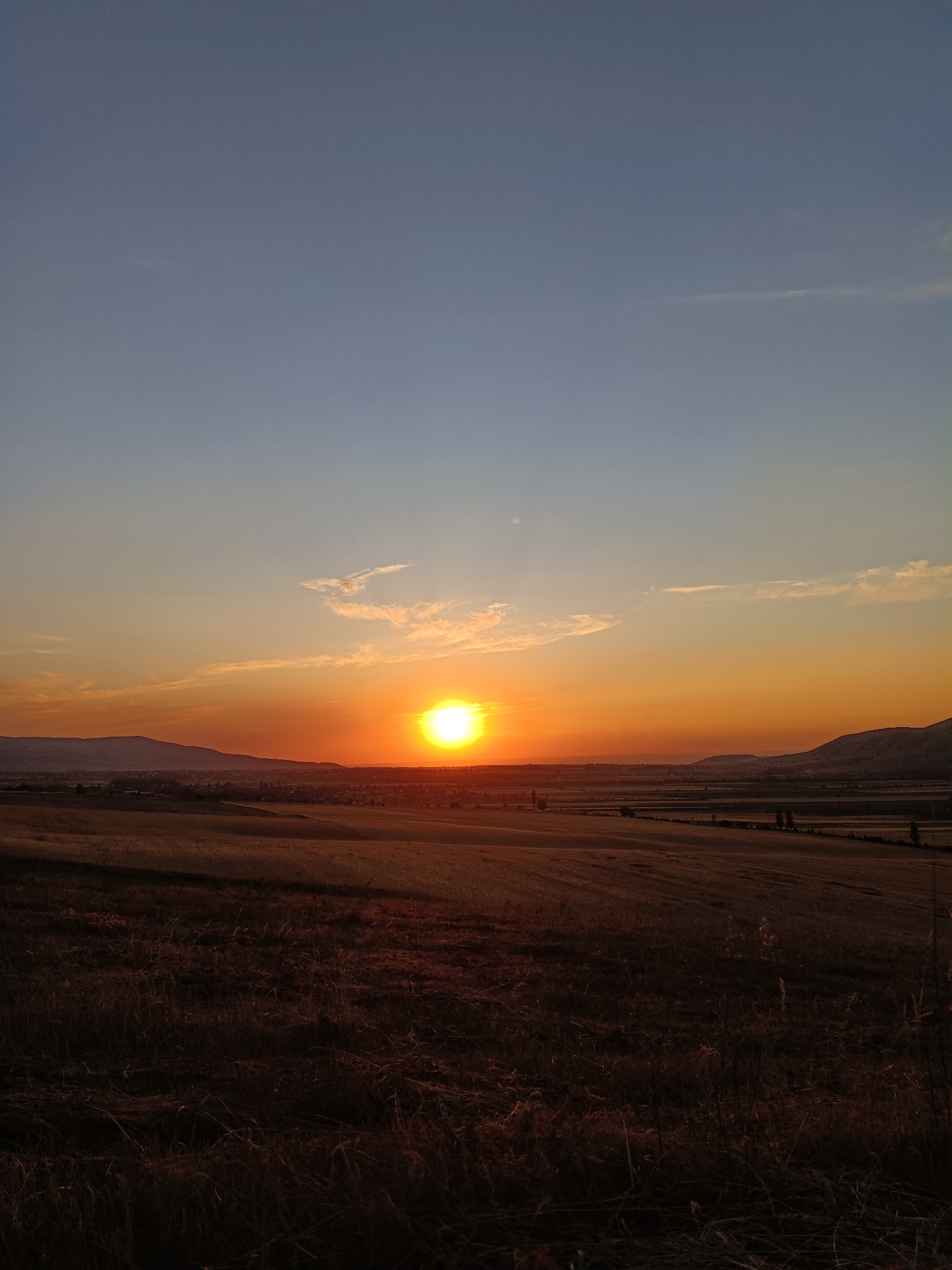 The image depicts a sunset over a rural landscape in Arashan, Kyrgyzstan. The sun, a bright orange orb, is positioned low on the horizon, casting a warm glow across the sky and illuminating a scattered cloud. The sky transitions from a pale blue at the top to shades of orange and yellow near the horizon. In the middle ground, rolling hills are visible, with a village or town nestled in the valley between them. The foreground is dominated by dry, reddish-brown grass and some sparse vegetation, suggesting a harvested field or uncultivated land. The overall scene conveys a sense of tranquility and natural beauty during the golden hour.