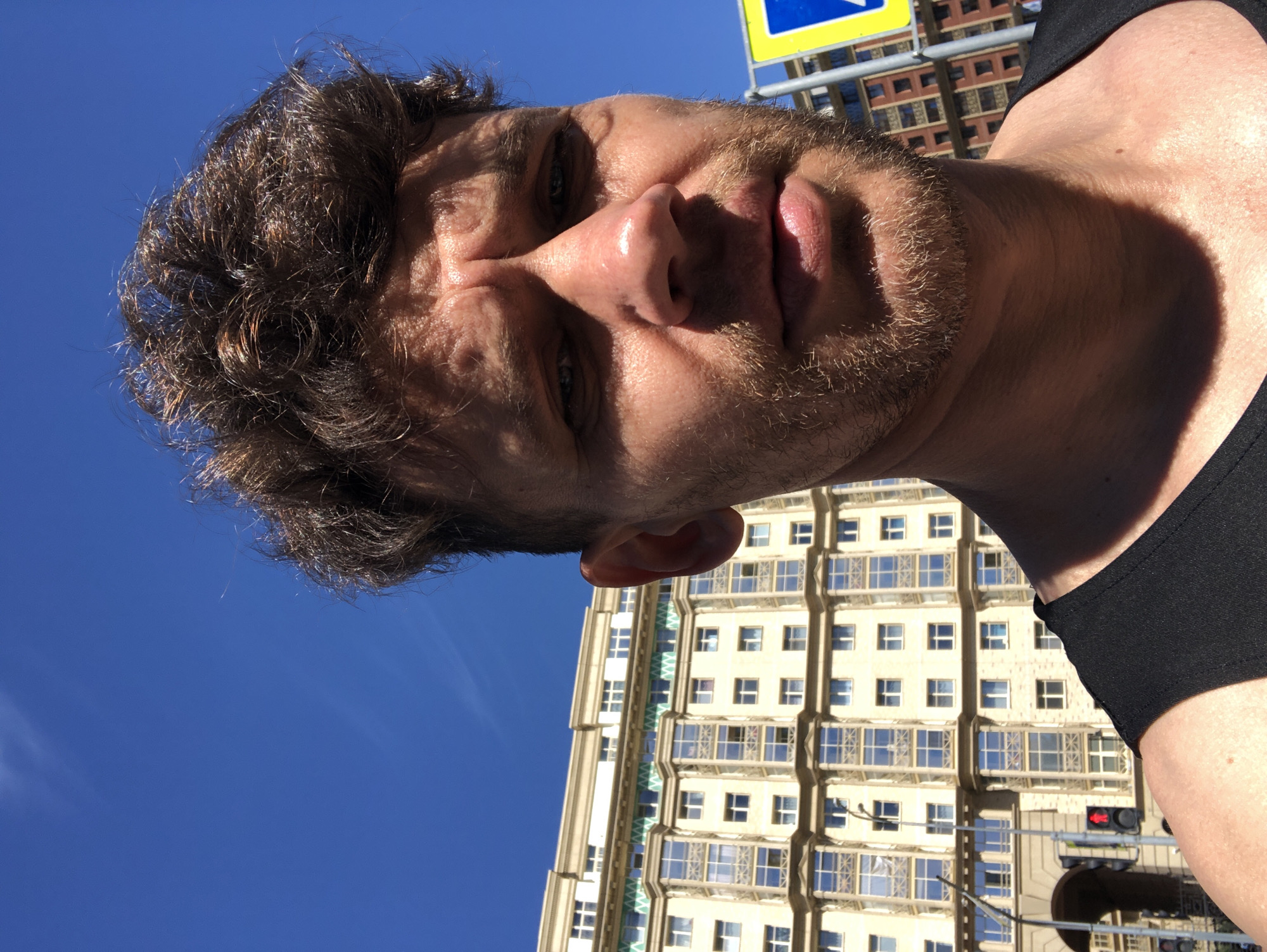 A man with messy brown hair and a light beard is taking a selfie outdoors. He is wearing a black tank top. Behind him, a large, cream-colored building with many windows and decorative elements is visible under a bright blue sky with a few thin clouds. To the right of the man, a yellow and blue street sign is partially visible. In the background, traffic lights and what appear to be more buildings can be seen. The overall impression is of a sunny day in an urban environment. The architecture suggests a location in Saint Petersburg, Russia.