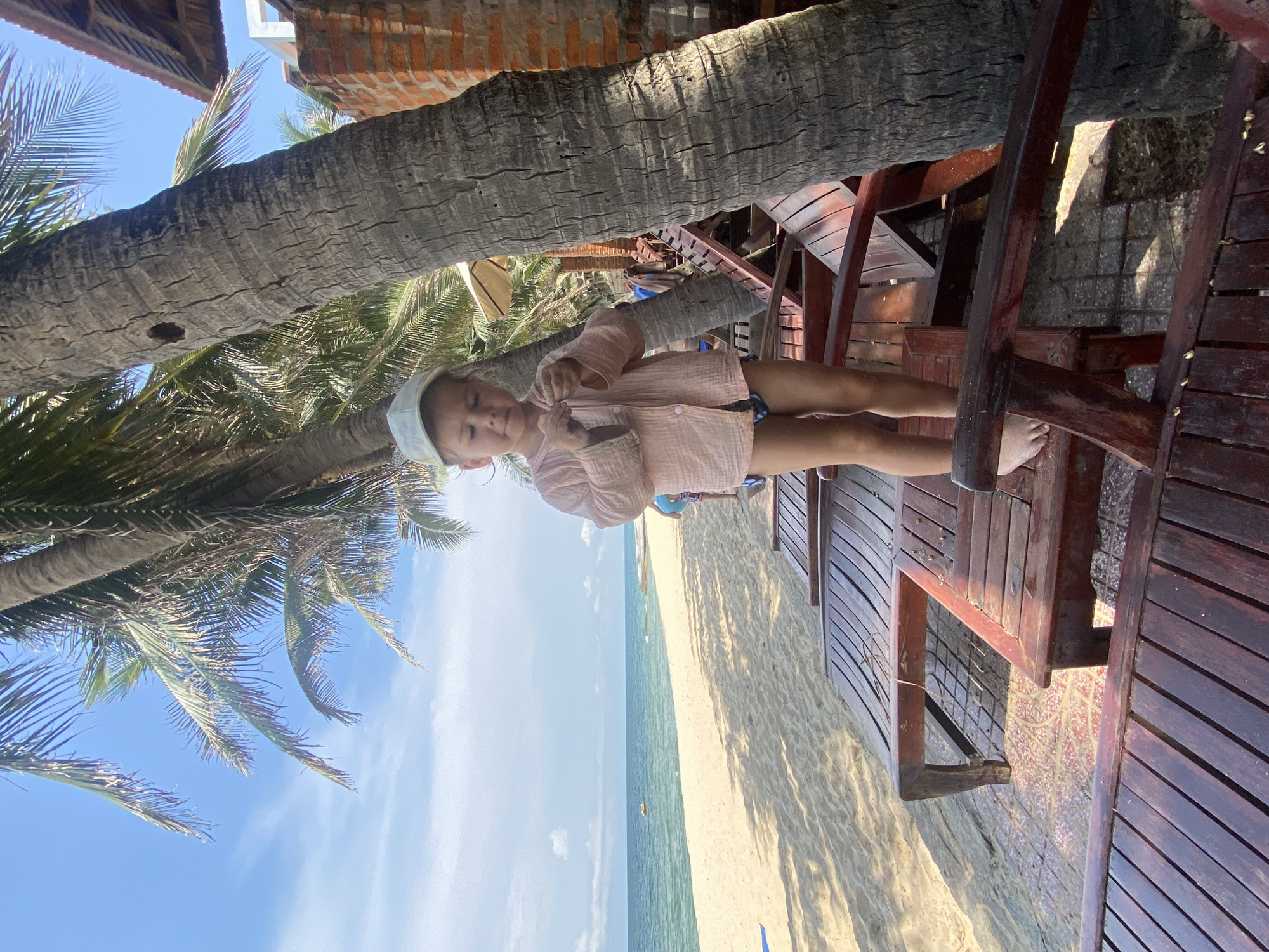 A young child wearing a light pink shirt, dark swim trunks, and a light-colored cap stands on a wooden lounger on a sandy beach. The child is looking down and appears to be holding something small in their hands. Behind the child, palm trees with their fronds visible against a blue sky suggest a tropical or subtropical location. The beach has a wide expanse of sand leading to the ocean, with a calm body of water visible in the distance. The scene is set in Phu Thuy Ward, Vietnam, indicated by the presence of palm trees and a beach environment. Several other wooden loungers are positioned on the sand, some of which are partially visible in the foreground. A brick wall is visible on the right side of the frame, suggesting the presence of a building.