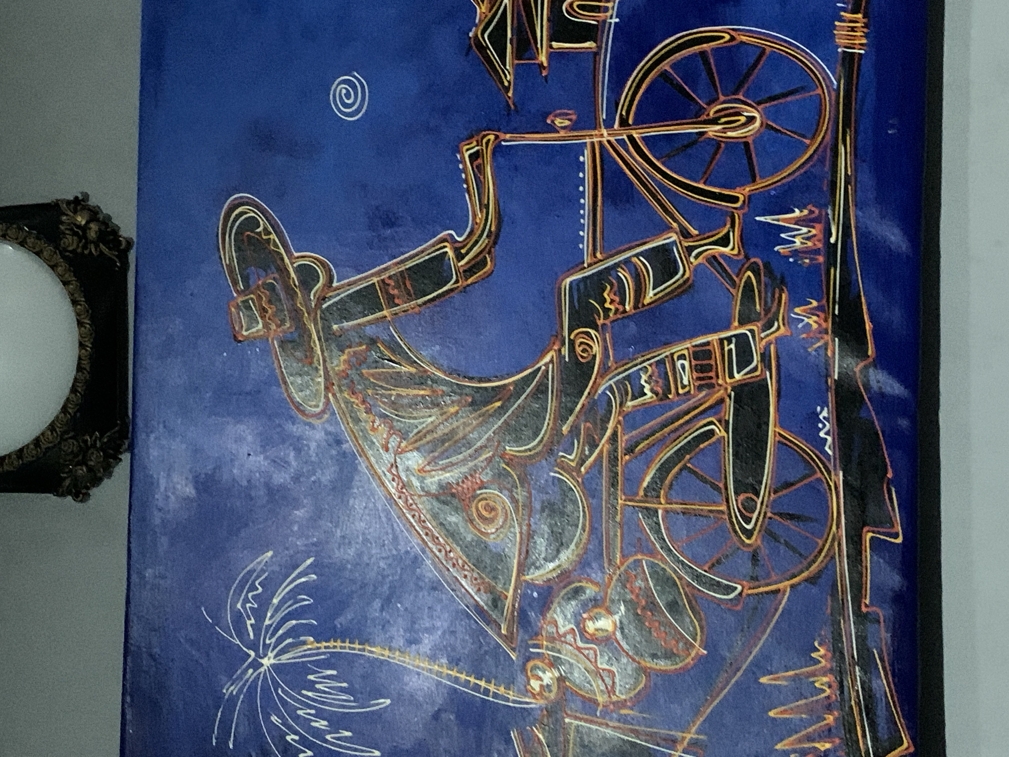 A painting with a blue background is partially visible next to a round mirror with an ornate black frame. The painting features abstract, stylized figures and objects outlined in gold, black, and white. Key elements include shapes suggestive of musical instruments, wheels, and figures that appear to be dancers or performers. A spiral symbol is in the upper-left. At the bottom of the image is a simplified, white drawing that resembles a palm tree. The location is Ibasa, Nigeria.