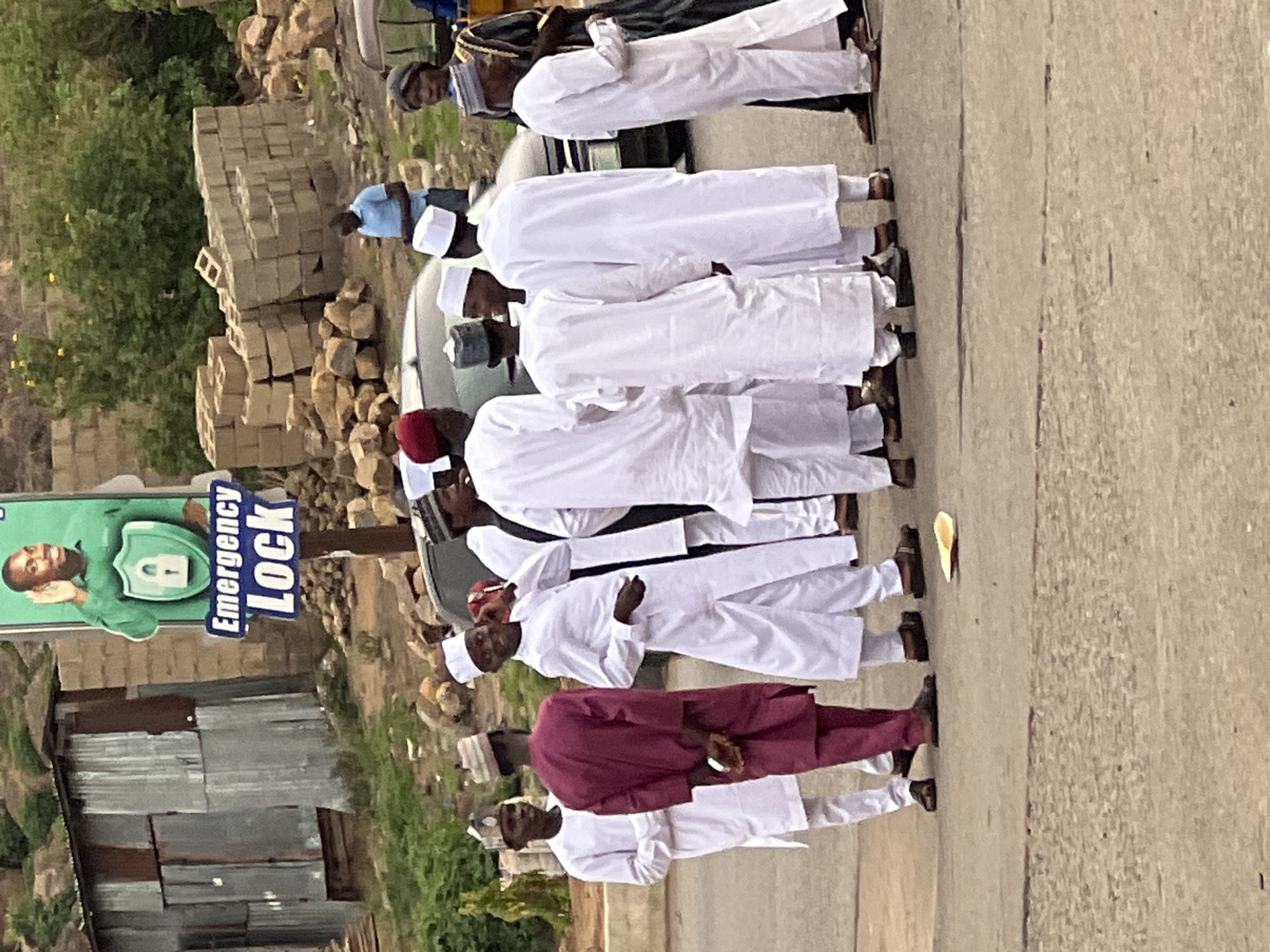 A street scene in Gura Top, Nigeria shows a line of at least 10 men wearing white traditional clothing and skullcaps walking on a paved road. One man wears a maroon outfit. The men are arranged in a single file line. In the background are buildings constructed of corrugated metal and concrete blocks. An "Emergency Lock" sign with a man's image is also visible.
