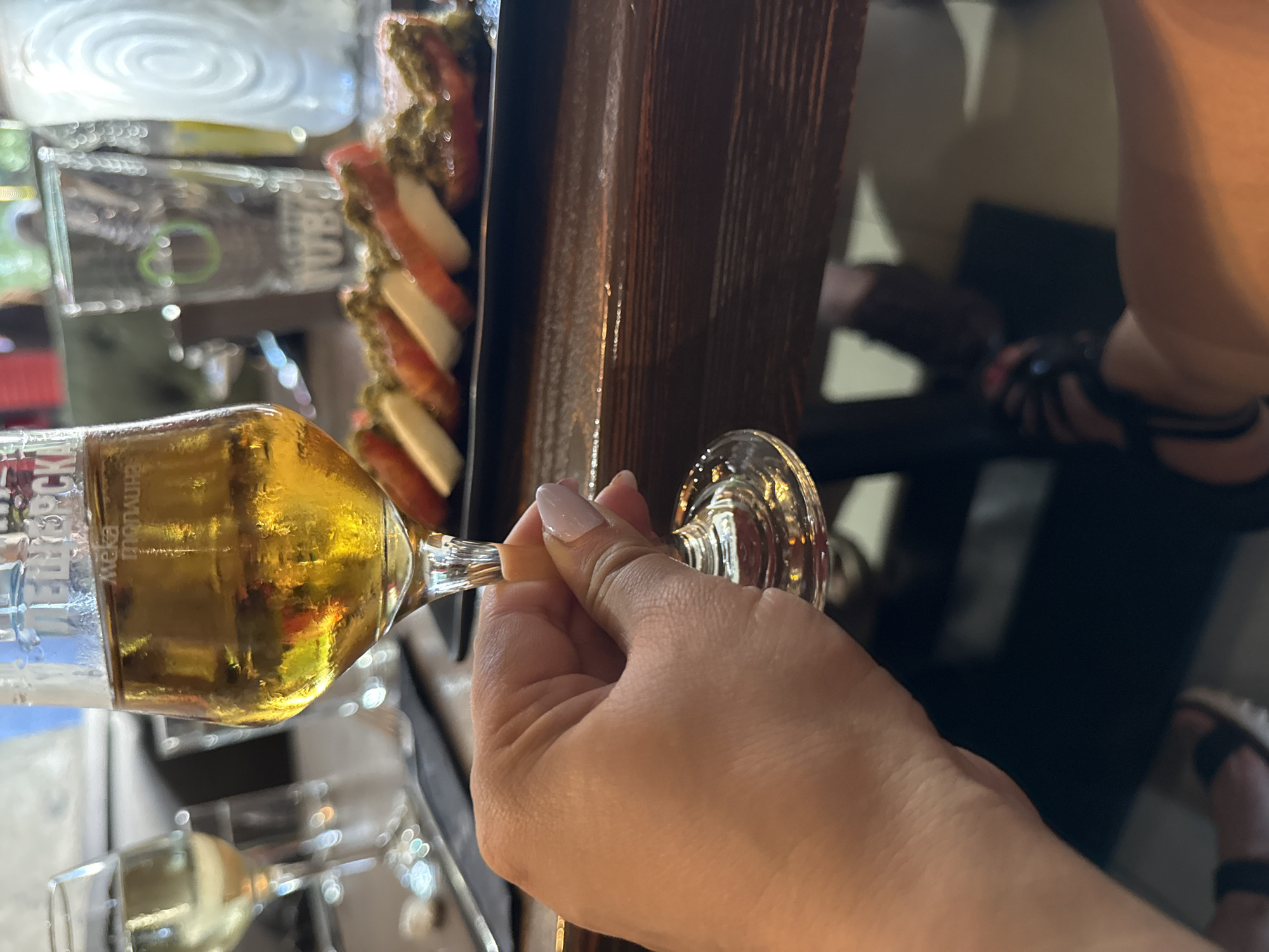 A first-person point-of-view image depicts a light-skinned hand holding a stemmed glass containing a golden-yellow, effervescent liquid, likely beer or an amber ale. Visible on the glass are partial Cyrillic characters, including "ПЕТЕРБУРГСК" and "МЕКА СТОЛИЦА". The hand has manicured fingernails with light pink polish. The setting is indoors, with a dark wooden table serving as the primary surface. In the background, out of focus, a dish of sliced tomatoes and mozzarella cheese topped with a green pesto-like substance is arranged on a black rectangular plate. Further in the background, another glass of lighter liquid, possibly white wine, is visible to the left, and various out-of-focus clear drinking glasses, one with a green logo, are visible to the right. A person's lower leg and foot, wearing dark sandals with a white sole, are partially visible in the bottom right corner beneath the table. The location is Unknown, Unknown.