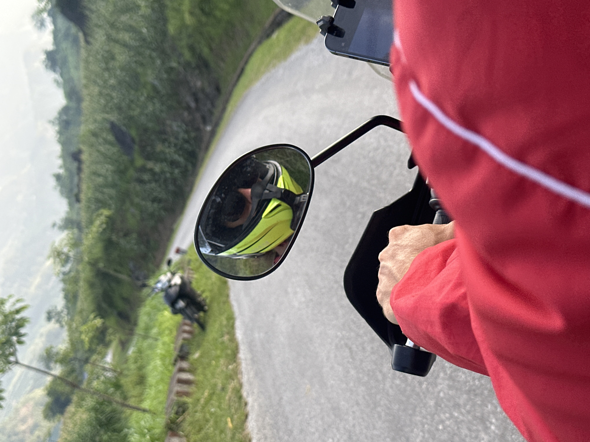 The image, taken from the perspective of a motorcyclist on a gray asphalt road, depicts a motorcyclist wearing a red jacket with white trim holding the handlebars. The side mirror reflects a rider wearing a yellow helmet, and a second motorcycle is visible down the road. The roadside shows green vegetation and a hillside. The scene appears to be in a mountainous region, consistent with the Du Già Commune, Vietnam.