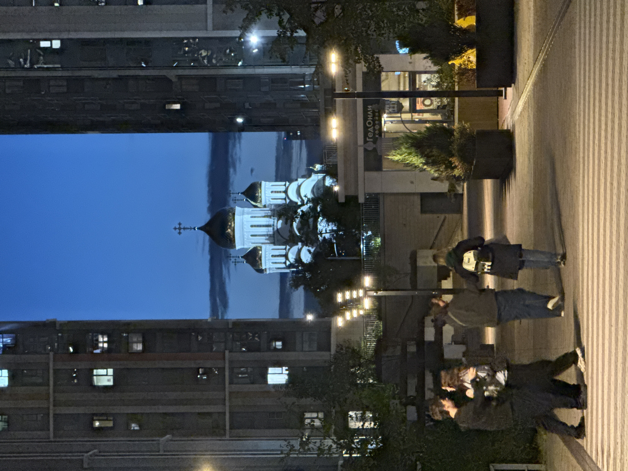 The image captures an outdoor scene at dusk in Moscow, Russia, likely in a modern urban development. The focal point is a white Orthodox church with golden domes, illuminated against a deep blue sky. The church is positioned between two tall, modern residential buildings. In the foreground, four individuals are walking across a pedestrian crossing. Two young people, a male and a female, are in the process of embracing, with the male’s arm around the female’s waist. Behind them, another couple, appearing to be older, walks in the same direction. The environment includes streetlights casting a warm glow on the pathway and planters with manicured evergreen trees. The pedestrian crossing is clearly delineated with white stripes. To the right, a commercial establishment with signage indicating "ГедОНли" and "кофейне" is visible. The overall atmosphere suggests a blend of contemporary urban architecture and traditional religious iconography, typical of many areas in Moscow.