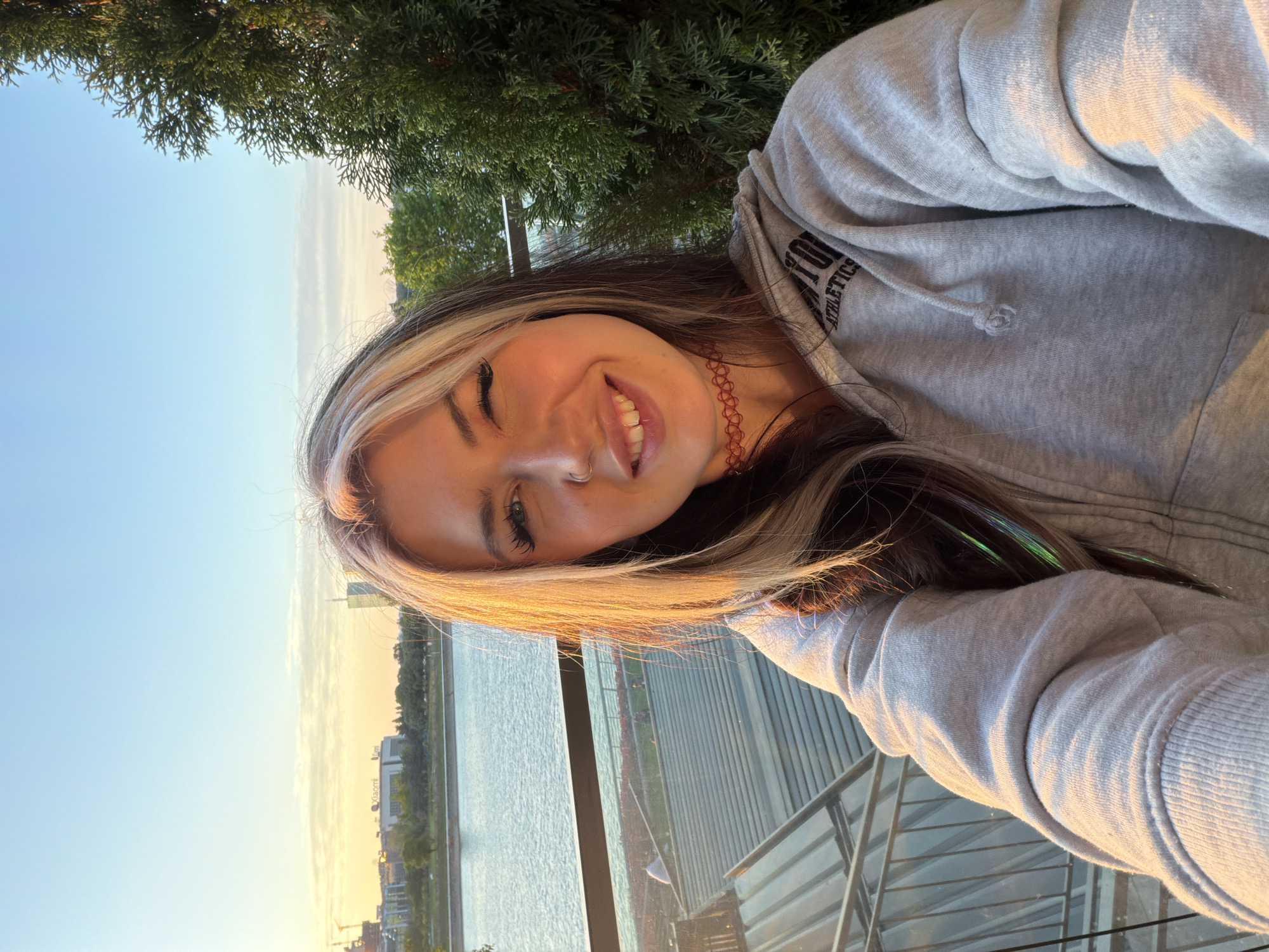 A young woman takes a selfie outdoors. She is smiling and wearing a grey hooded sweatshirt and a red choker. Her hair is dark with blonde highlights. The background features a river, buildings, and trees. The location is Belgrade, Serbia.