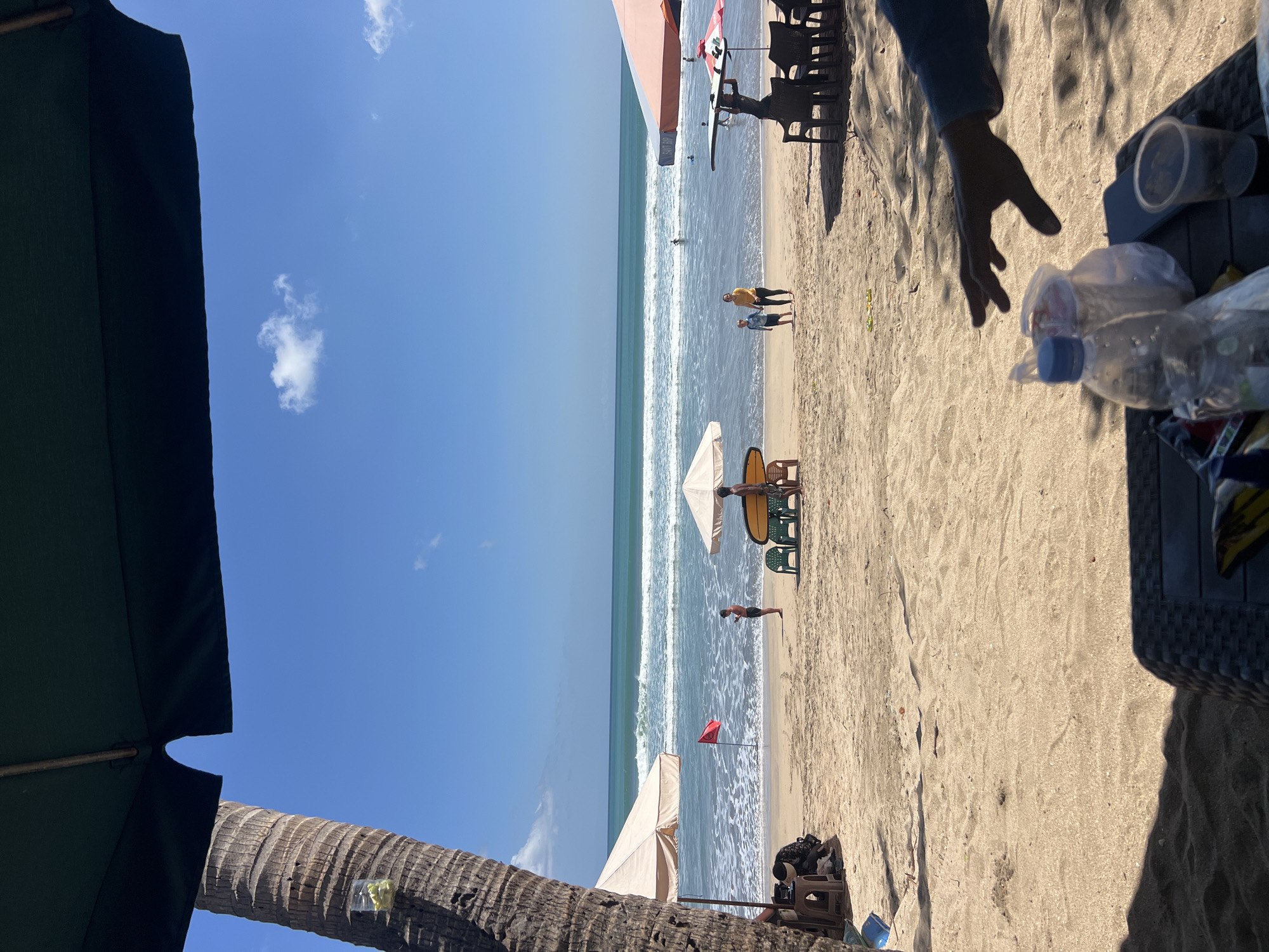 The image depicts a tropical beach scene in Kuta, Indonesia, under a clear blue sky. In the foreground, a person's hand is reaching out over a table with drinks and plastic bags. The beach is sandy, with several beach umbrellas providing shade. A palm tree trunk is visible on the left side of the image. In the middle ground, people are seen walking on the sand and near the water. Waves are breaking on the shore, and the ocean stretches out to the horizon. A red flag is planted in the sand. In the background, more people are visible in the water.