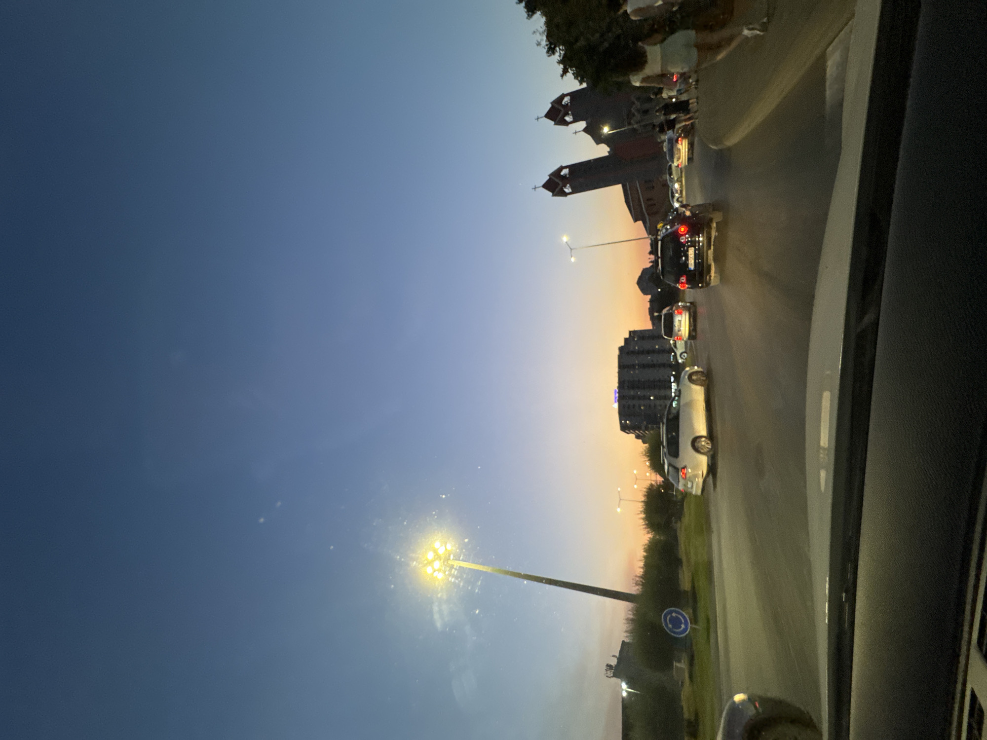 The image depicts a street scene at dusk in Bjeliši, Montenegro. The sky transitions from a deep blue at the top to hues of orange and yellow near the horizon. In the foreground, the dashboard of a vehicle is visible, suggesting the photo was taken from a car. The street is occupied by several cars, some in motion and others parked. A prominent street lamp with multiple bright bulbs illuminates the left side of the scene. In the background, buildings and trees are discernible. To the right, a large, distinctive church with two towers and crosses on top is visible. Several people are walking on the sidewalk near the church. The overall atmosphere is that of a busy urban environment at the end of the day.