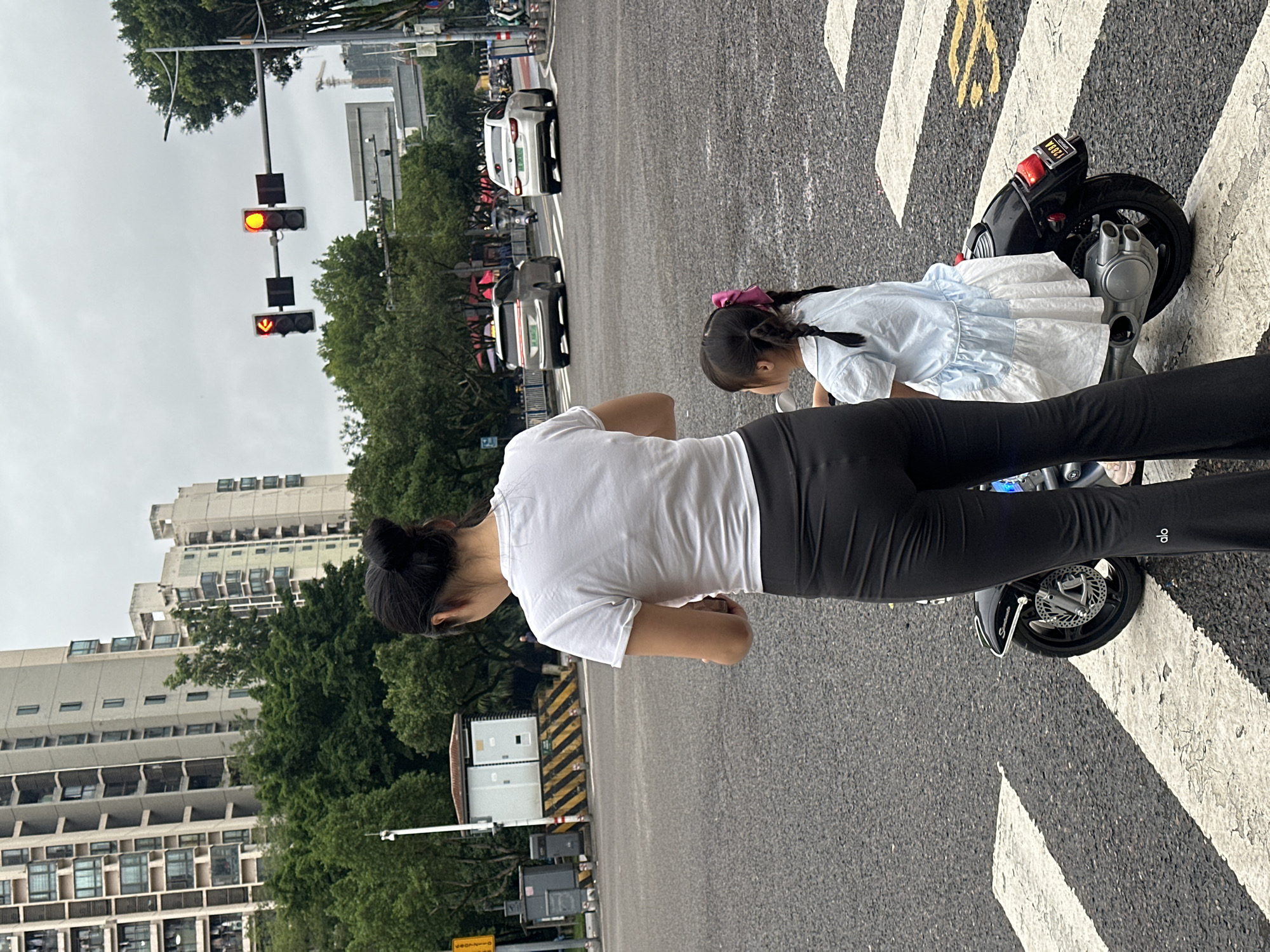 The image shows two individuals, a woman and a young girl, on a street. The woman is wearing a white t-shirt and black leggings, with her hair pulled back in a bun. The girl is wearing a light blue dress and has her hair in braids. They are standing next to a small, black electric scooter with a license plate that reads "26B5K". The scene appears to be a street crossing with white and yellow striped pedestrian markings.
In the background, there are cars on the road and buildings on either side. Traffic lights are visible, with one displaying a yellow light and another a red arrow. The sky is overcast, suggesting a cloudy day. The architecture of the buildings, particularly the multi-story structures with many windows, hints at an urban environment. The overall impression is a moment captured on a city street, possibly waiting to cross or taking a break. The young girl seems to be looking down, while the woman is facing away from the camera. There is no discernible text on the people or the scooter, other than the license plate. The location context provided, Chongqing High-tech Zone, China, helps to situate the scene within a specific geographical area known for its technological development and urban landscape.