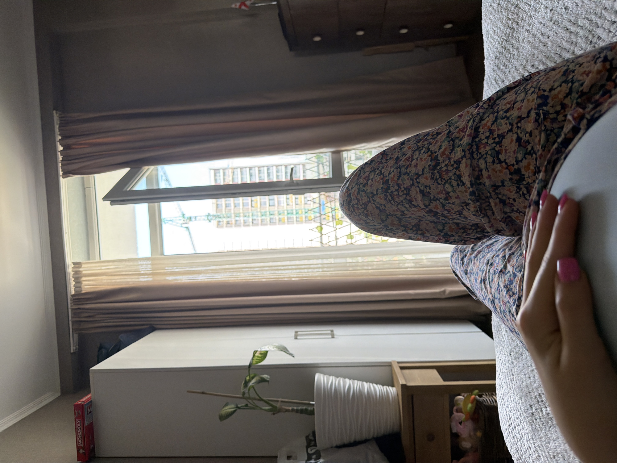 A person, seen from the waist down, is sitting on a sofa in what appears to be a bedroom. They are wearing a floral-patterned dress. The scene is viewed from a low angle, with the camera tilted sideways. Through a window, we see a cityscape with buildings under construction, suggesting an urban environment. The time of day is difficult to determine, but the light suggests it could be daytime. The overall mood is relaxed and casual.
Notable details include a white wardrobe in the foreground, a Monopoly box on the floor next to it, and a potted plant on top of the wardrobe. A small wooden shelf unit with various items is also visible. The window is partially open, and the curtains are drawn back. The exterior view reveals construction cranes, indicating ongoing development in the area. The cropped images help to focus on specific elements, such as the person's dress and hands, the details of the window and curtains, and the furniture and objects in the room. The location context is identified as Tbilisi, Georgia.