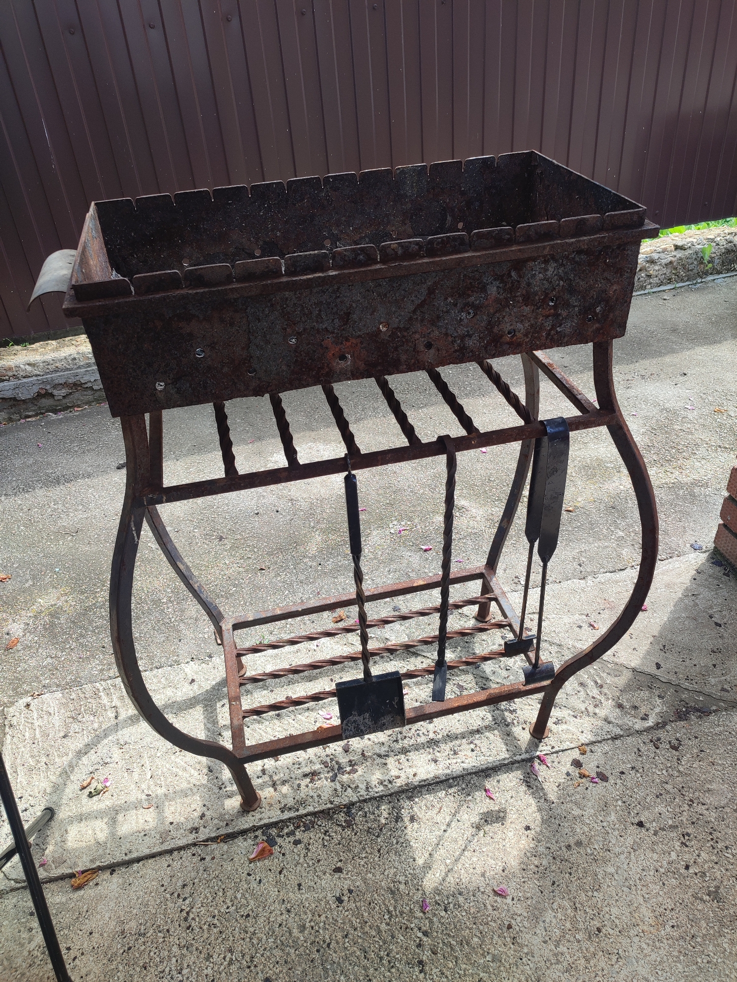 An outdoor scene in Kumertau, Russia, shows a rusted metal barbecue grill on a concrete surface. The grill is rectangular with an open top and a stand made of decorative wrought iron legs. Attached to the stand are three barbecue tools: a shovel, a fork, and a poker. The grill is positioned in front of a brown corrugated metal fence. The ground is littered with small debris, including fallen leaves and petals. The lighting suggests it is daytime, with shadows cast on the concrete. No people are visible in the image.