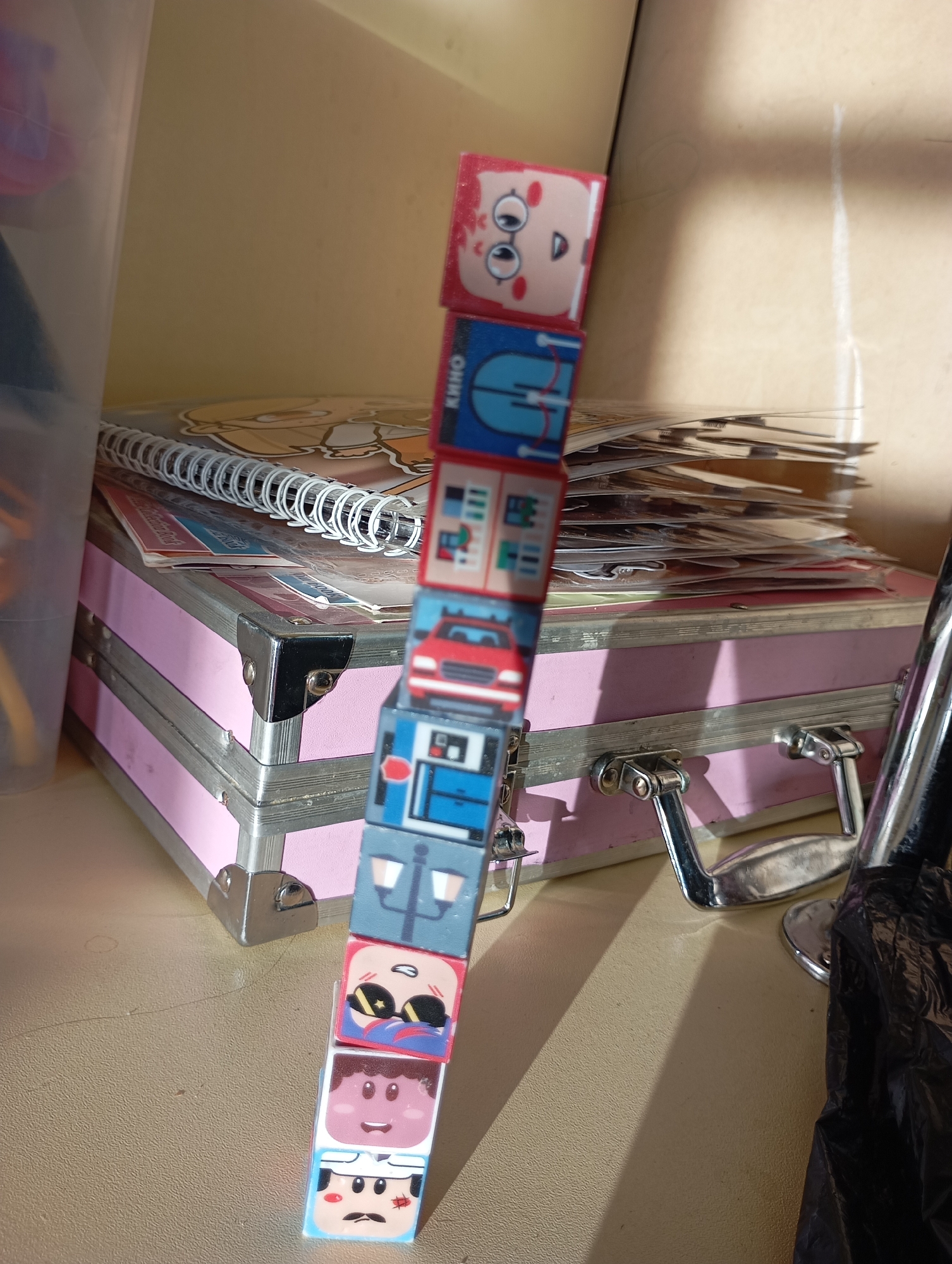 A stack of eight colorful building blocks with cartoon faces is standing on a tan surface in a cluttered indoor environment. The blocks are slightly tilted and appear to be balancing precariously. Behind the blocks, there is a pink and silver metal suitcase-style box with a pile of papers, a notebook, and other items on top. In the background, there is a light-colored wall and a clear plastic container. The scene is brightly lit.