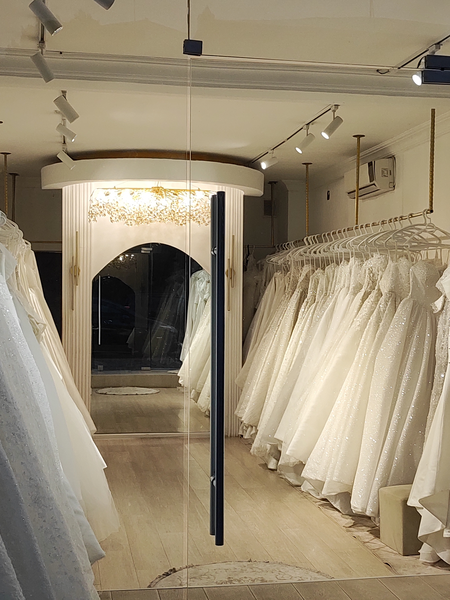 A wedding dress shop is depicted from the exterior, looking through a glass door. The shop interior is brightly lit by a large, ornate chandelier and several track lights on the ceiling. Rows of white wedding dresses are displayed on racks, with some dresses hanging in the foreground. The floor appears to be made of light-colored wood. To the right, an air conditioning unit is visible on the wall. The shop has an arched doorway leading to a darker area within.