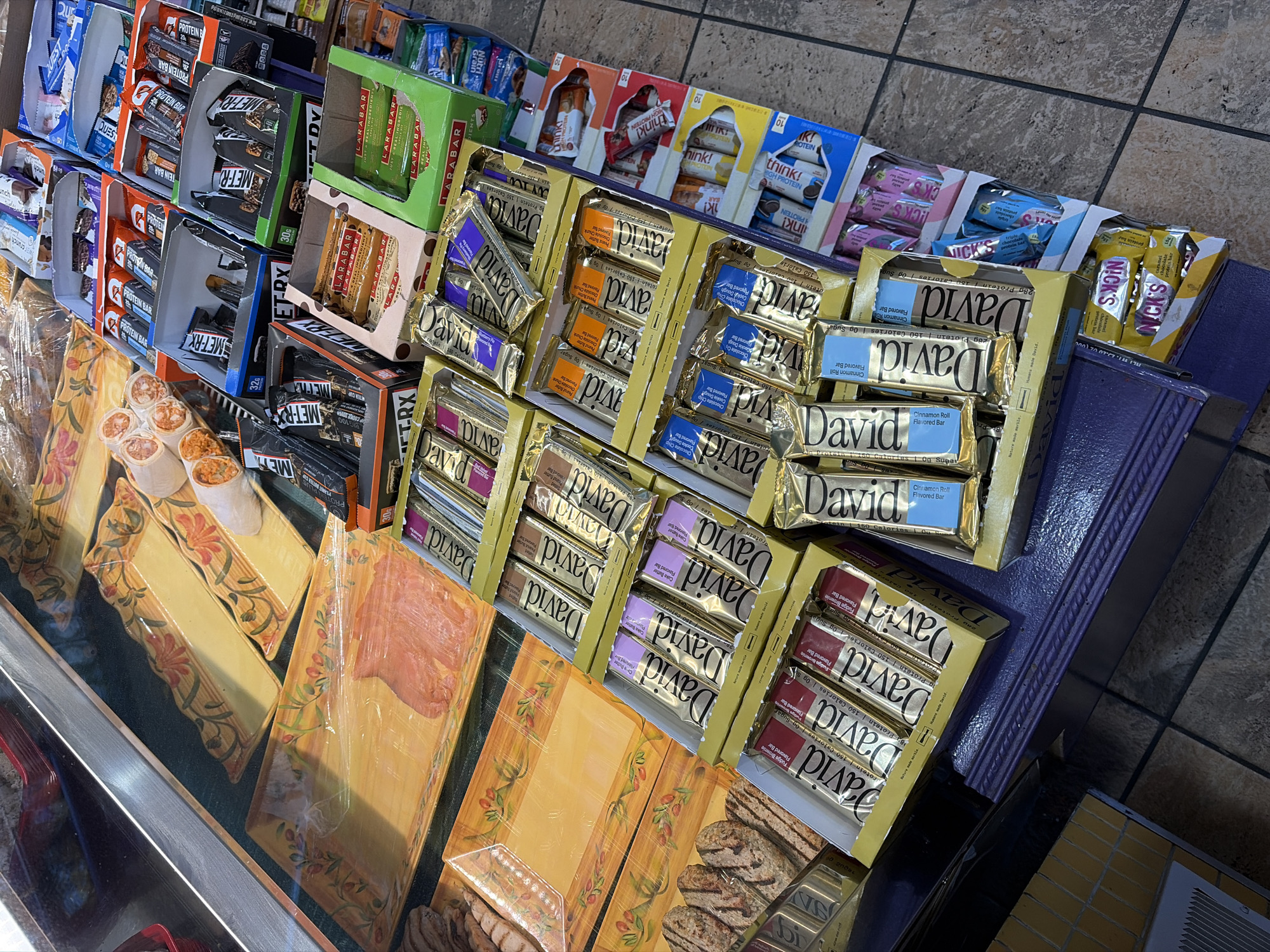 This is a high-angle, indoor shot of a convenience store display filled with various protein and snack bars. The bars are neatly arranged in their original packaging on top of yellow patterned trays. Prominently featured is "David" protein bars with different flavors indicated by color-coded labels: Cinnamon Roll, Chocolate Chip Cookie Dough, Cake Batter, and Fudge Brownie. Other brands include "MET-Rx", "Larabars", "Think", "Quest" and "Nicks". The scene is on a countertop made of a dark gray material, with a tiled floor surrounding the display. The scene doesn't have any people, or interactions with them. The location in New York City is undetermined.