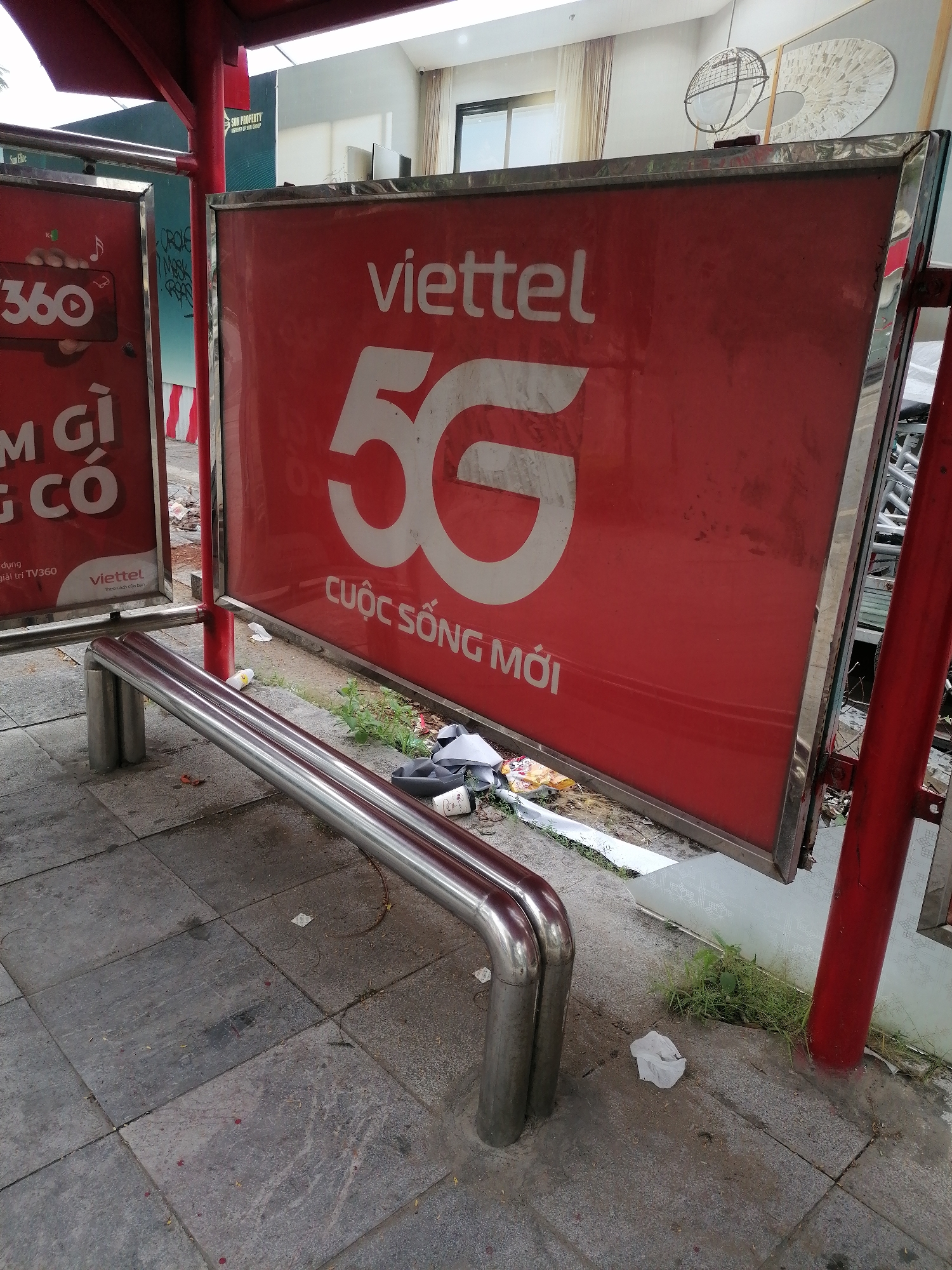 The image shows an outdoor advertising display in Bai Chay Ward, Vietnam. The prominent ad features the "viettel" logo and "5G CUOC SONG MOI". Another advertising panel partially visible on the left displays "360" and Vietnamese text. A metal bench is positioned in front of the ads. Debris and trash are scattered on the ground around the base of the structure. The ground consists of gray paving stones.