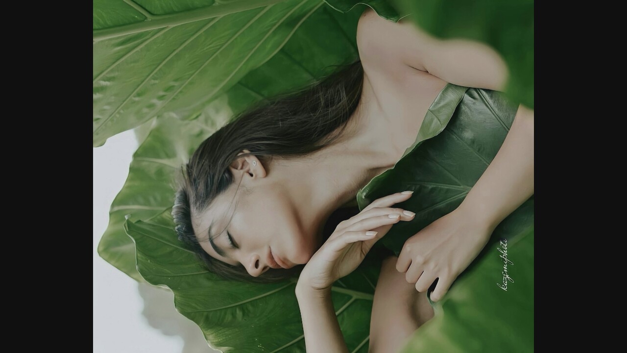 A woman with dark hair lies with her eyes closed, nestled among large green leaves. Her head is tilted back, and her right arm is bent, with her hand resting on her chest. A smaller leaf covers her upper chest and shoulder area. Her left arm is bent forward, with her hand resting on another large leaf. A watermark reading "kazemiphoto" is visible in the lower left corner of the image. The overall setting appears to be natural and lush.