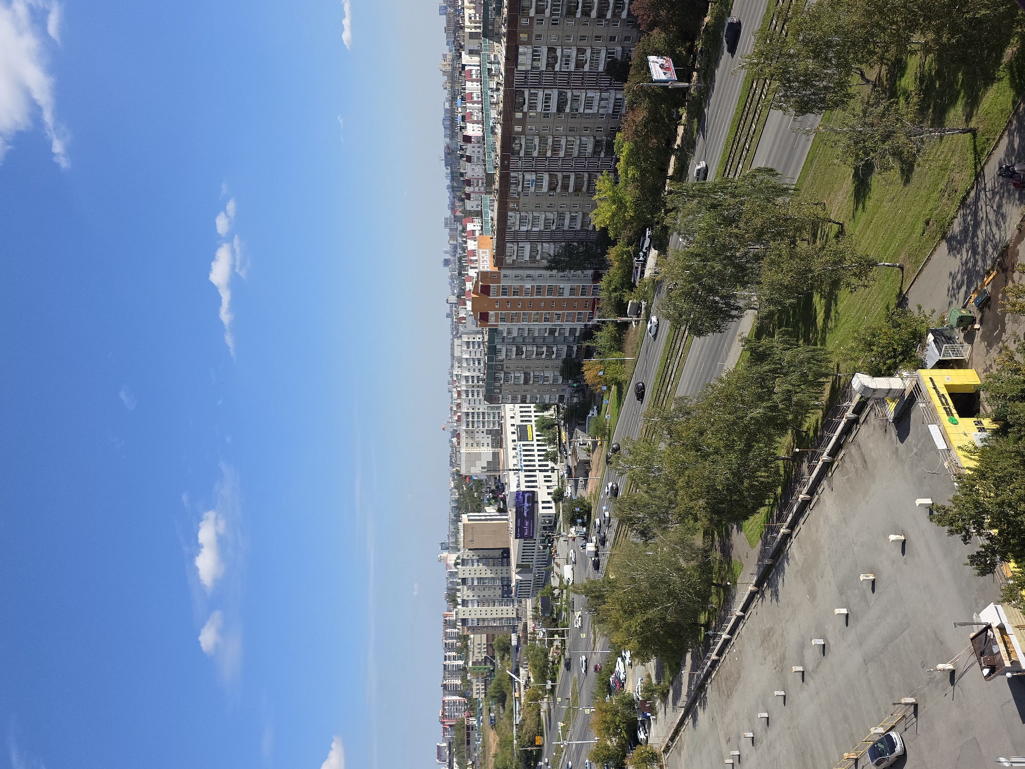 This is an aerial, eye-level shot of a city scene in Chelyabinsk, Russia, under a bright, partly cloudy blue sky. The foreground features a large, gray, paved parking lot with several white, rectangular bollards evenly spaced. A silver car is parked on the far left of the lot, and a yellow building with a green entrance is visible on the lower right. Beyond the parking lot is a green, grassy slope interspersed with trees, leading down to a multi-lane road carrying moderate traffic, including cars and a white van. There are also tram tracks running parallel to the road.
The middle ground is dominated by a long stretch of apartment buildings, some with balconies and varying architectural styles, including a taller building with orange accents and a large banner. Another building in this area features a large advertisement billboard. Trees line the median of the road and parts of the sidewalk.
The background reveals a dense urban landscape with numerous residential buildings of different heights and colors, stretching as far as the horizon. The overall impression is of a sunny day in a developed urban area.