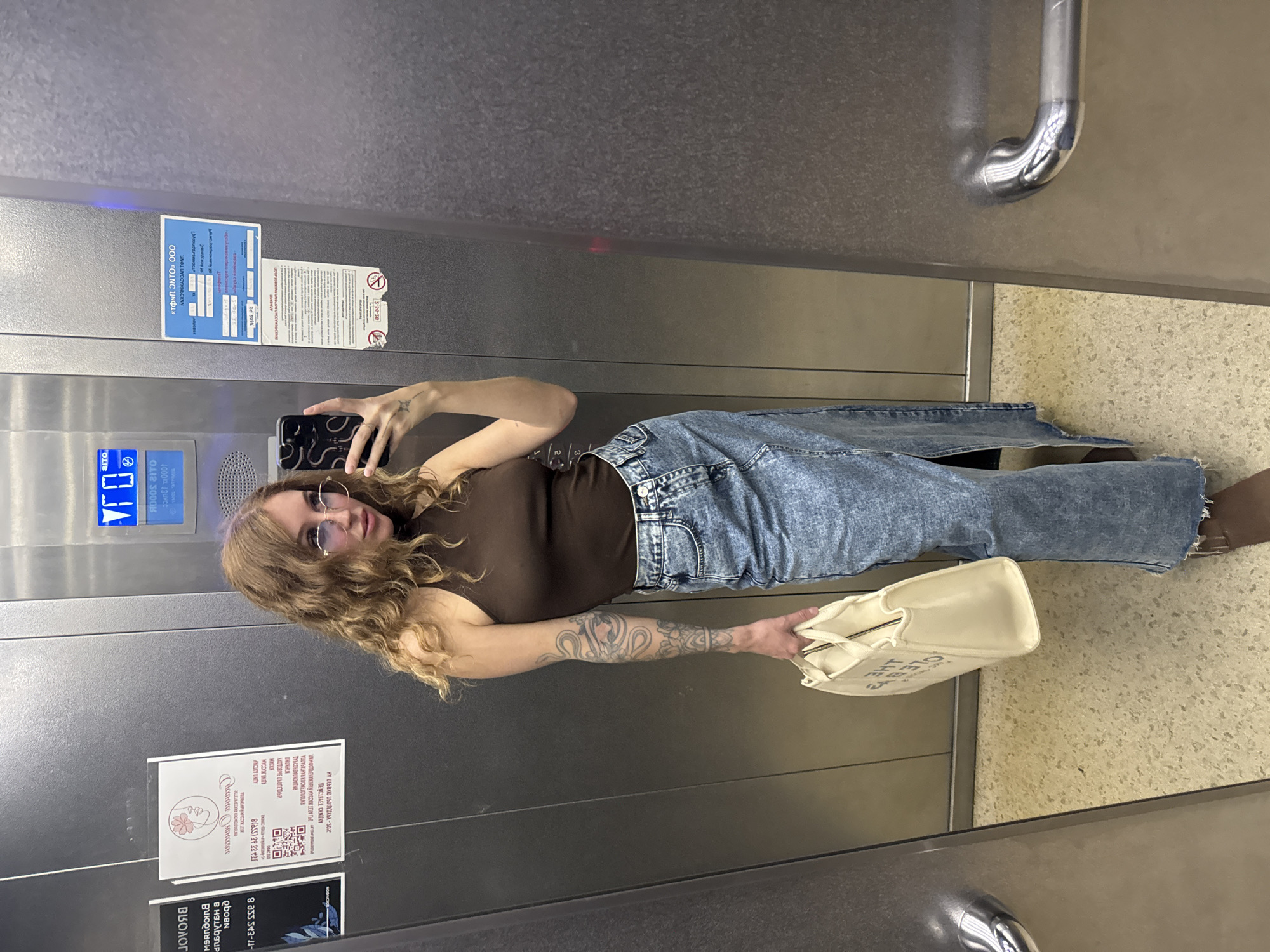 A full-body, front-facing mirror selfie is captured in an elevator. The subject is a woman with curly, light brown hair and light blue-tinted glasses. She is wearing a brown sleeveless turtleneck top and a long, light blue denim skirt with a high slit. She is holding a cream-colored tote bag in her left hand. Tattoos are visible on her left arm. The elevator's interior is metallic gray with a speckled beige floor. A metal handle is visible on the right side of the elevator. There are various informational posters on the elevator walls. One poster on the left features a stylized drawing of a woman's face with a flower. A poster on the right contains text in Cyrillic. The elevator panel with buttons is visible at the top of the frame. The overall environment appears to be a typical urban elevator. The location is identified as Chaikovsky, Russia.