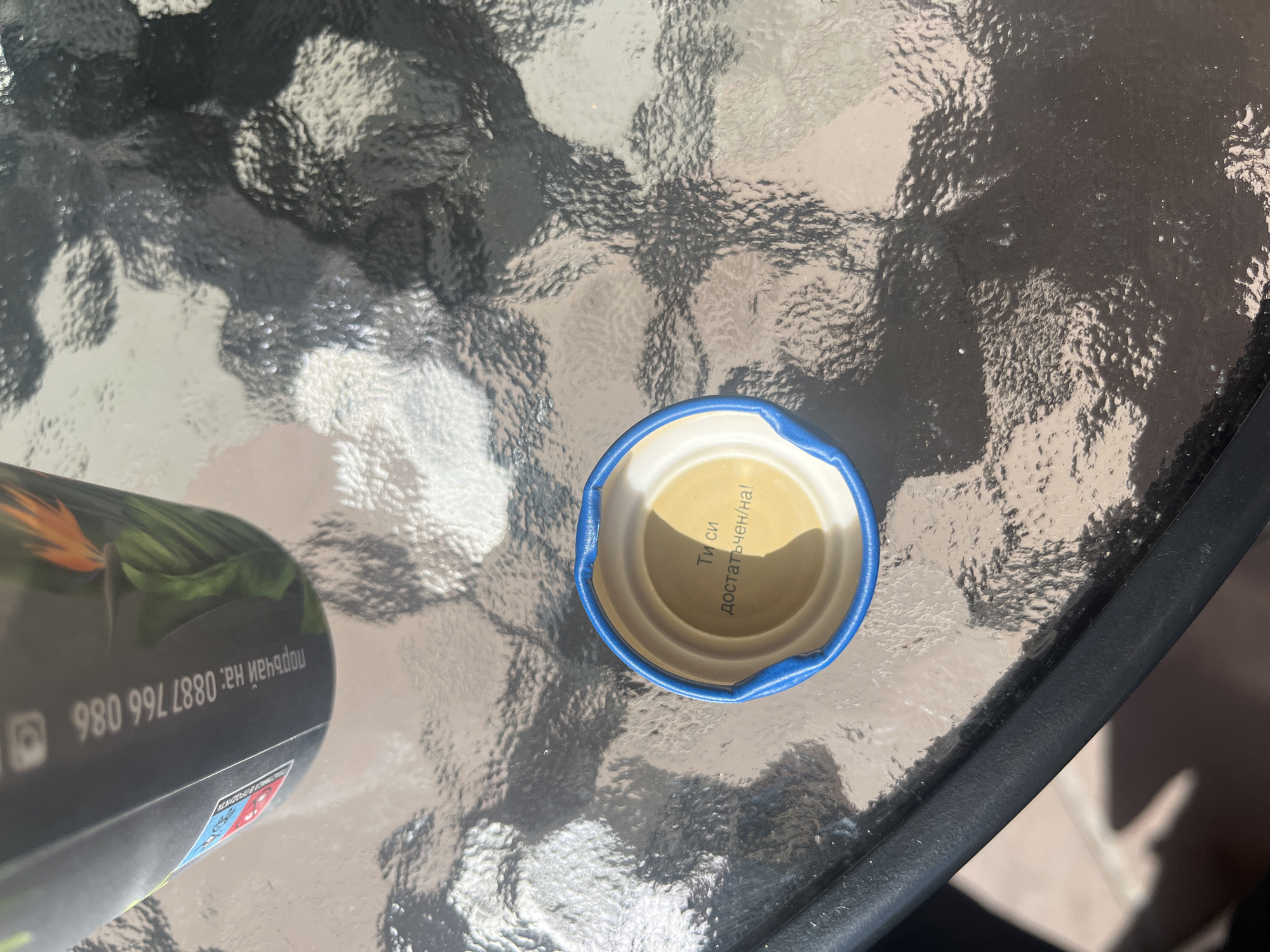 An overhead shot captures a blue and white bottle cap resting on a textured glass table, alongside part of a dark beverage can or bottle. The setting appears to be outdoors or a brightly lit area, indicated by harsh shadows and strong sunlight reflecting off the glass surface. The table's surface has a distinct, rippled texture, creating abstract patterns of light and shadow.
Inside the bottle cap, Cyrillic text is visible, angled slightly: "Ти си достатъчен/на!". This translates to "You are enough" (gender-neutral/feminine ending).
To the left, a portion of a dark-colored can or bottle is visible, featuring a tropical-themed illustration with green leaves and an orange flower. Upside down text on the can reads "поръчай на:" (meaning "order at:") followed by a phone number: "0887 766 980".
The overall impression is a casual outdoor scene, possibly a cafe or patio in Plovdiv, Bulgaria, given the Cyrillic text and provided location context. The time of day is clearly daytime due to the strong, direct sunlight.