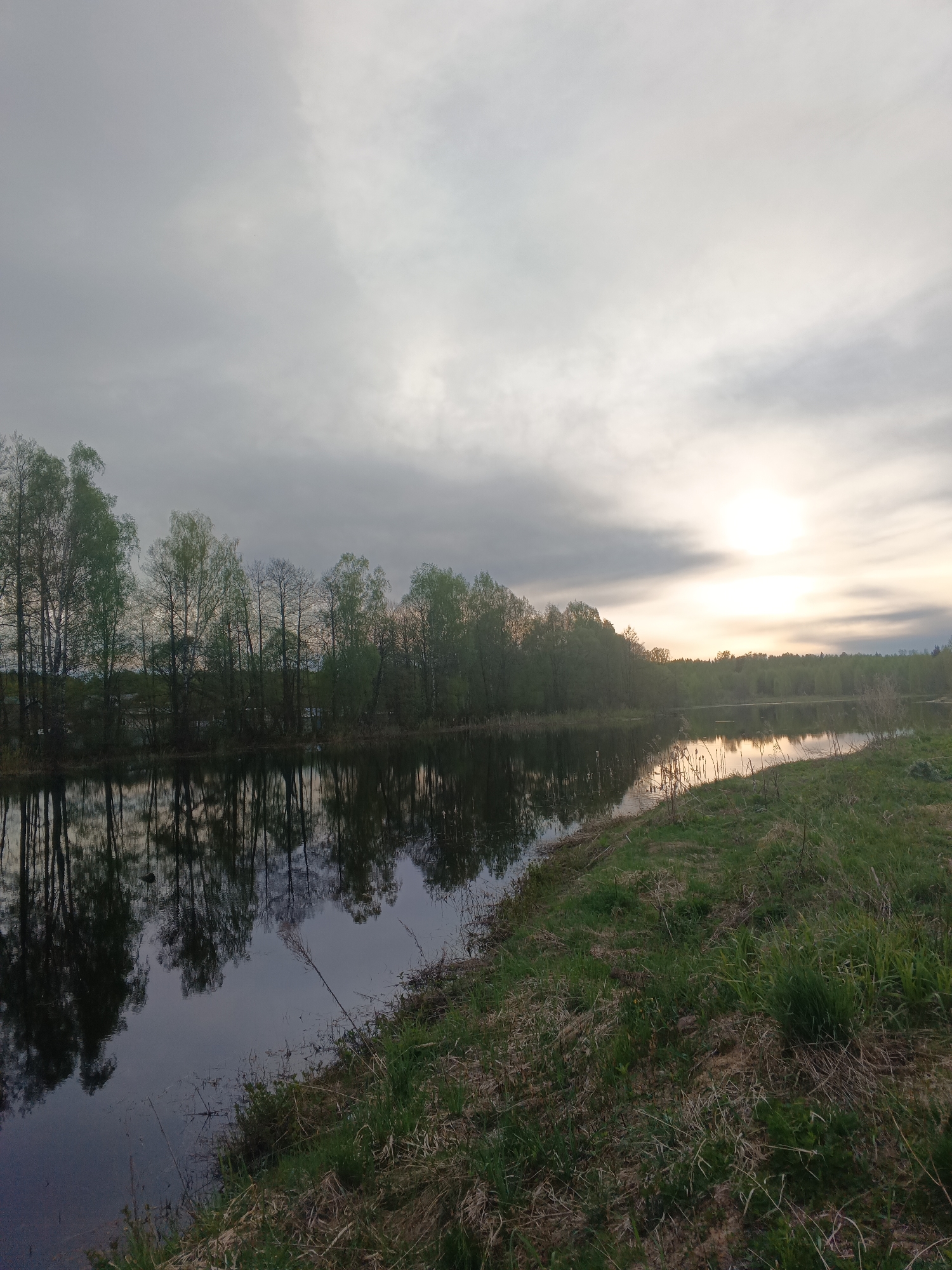 The image depicts a tranquil, rural scene in Красный Маяк, Russia. A narrow body of water, likely a river or canal, runs through the landscape, reflecting the silhouettes of trees lining its bank. The trees exhibit early spring foliage. The opposite bank is covered in dense grass and vegetation. The sky is overcast with low clouds, obscuring the sun, which casts a diffused light across the horizon. There are no people visible in the image.