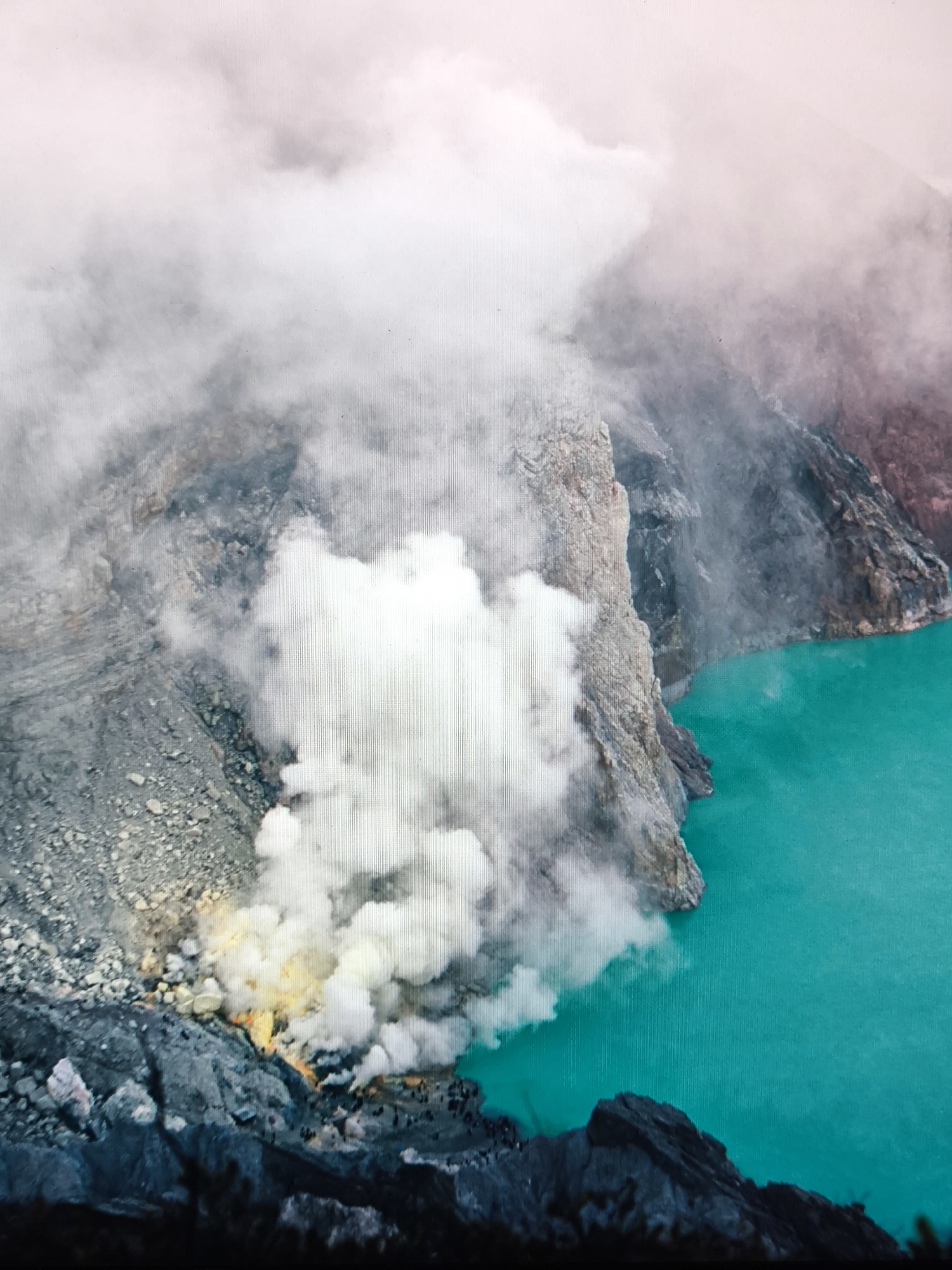 Location: Volcanic crater, specific city and country unknown.
The image captures an active volcanic crater scene. A large, irregularly shaped body of vivid turquoise water, consistent with a highly acidic crater lake, dominates the right and central-right portions of the frame. This lake is bordered by steep, jagged, dark grey and brown volcanic rock formations, which constitute the crater walls. On the left side of the image, the inner crater slopes display a rugged, rocky terrain composed of grey and reddish-brown volcanic material. Dense, opaque plumes of white and light grey smoke and gas are actively emanating from multiple vents positioned at the base of the crater wall on the left, immediately adjacent to the lake. A distinct yellow flame or glow is visible amidst these emissions, indicative of active sulfur combustion. Several small, dark figures, identified as human individuals, are present on the rocky ground near the sulfurous vents and plumes; approximately 10 to 15 individuals are discernible. The upper portion of the scene is occupied by a pale pink-grey sky, partially obscured by additional layers of diffused white and grey atmospheric or volcanic cloud cover.
