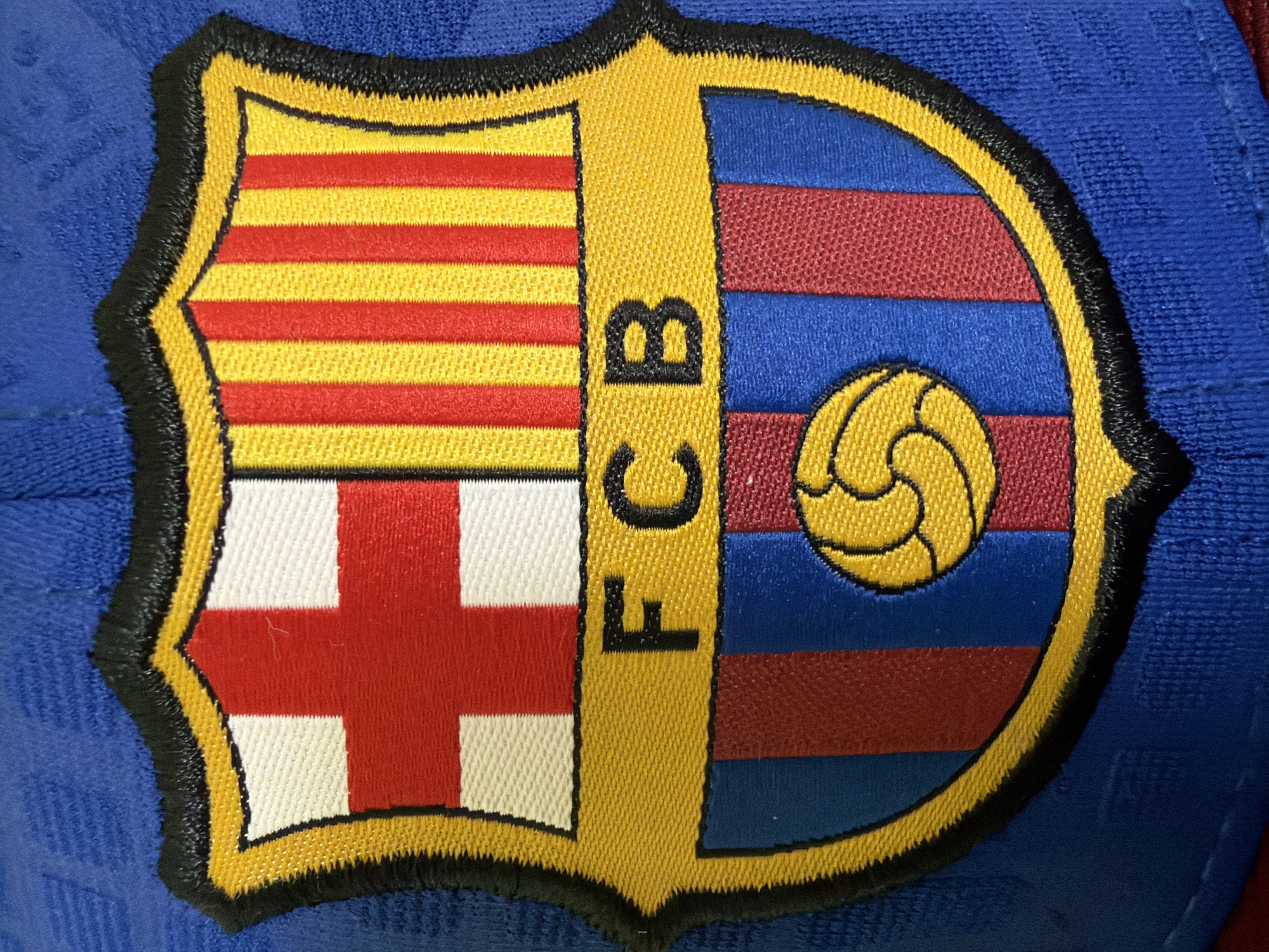 The image shows a close-up view of the crest of FC Barcelona, a Spanish professional football club. The crest is embroidered on a blue fabric, likely a jersey or hat. The crest features the Cross of Saint George and the Catalan flag in the top half, and vertical red and blue stripes in the bottom half. In the center of the bottom half, the letters "FCB" are embroidered in black. A yellow football with a black outline is positioned in the lower portion of the crest. The crest is outlined with yellow and black embroidery. The image does not provide any information about the location in the city of Kyiv, Ukraine, nor does it depict any people, actions, or interactions.