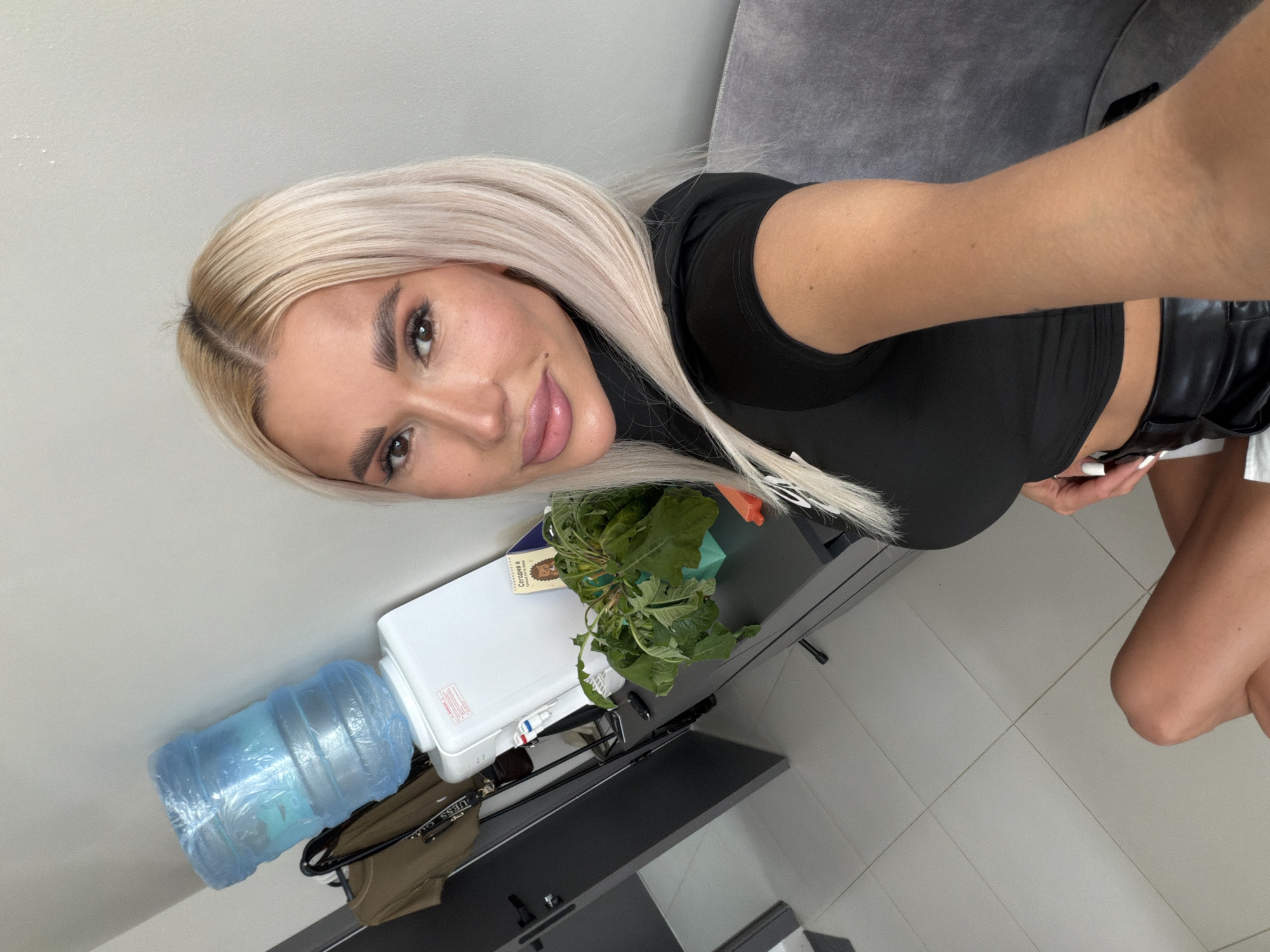 The image shows a woman with blonde hair taking a selfie. She is wearing a black top and leather shorts. She is sitting on a gray couch. In the background, there is a water cooler, a plant, and a door. The image appears to be taken indoors, in a room with tiled floors and light gray walls. The overall impression is of a casual, personal snapshot.