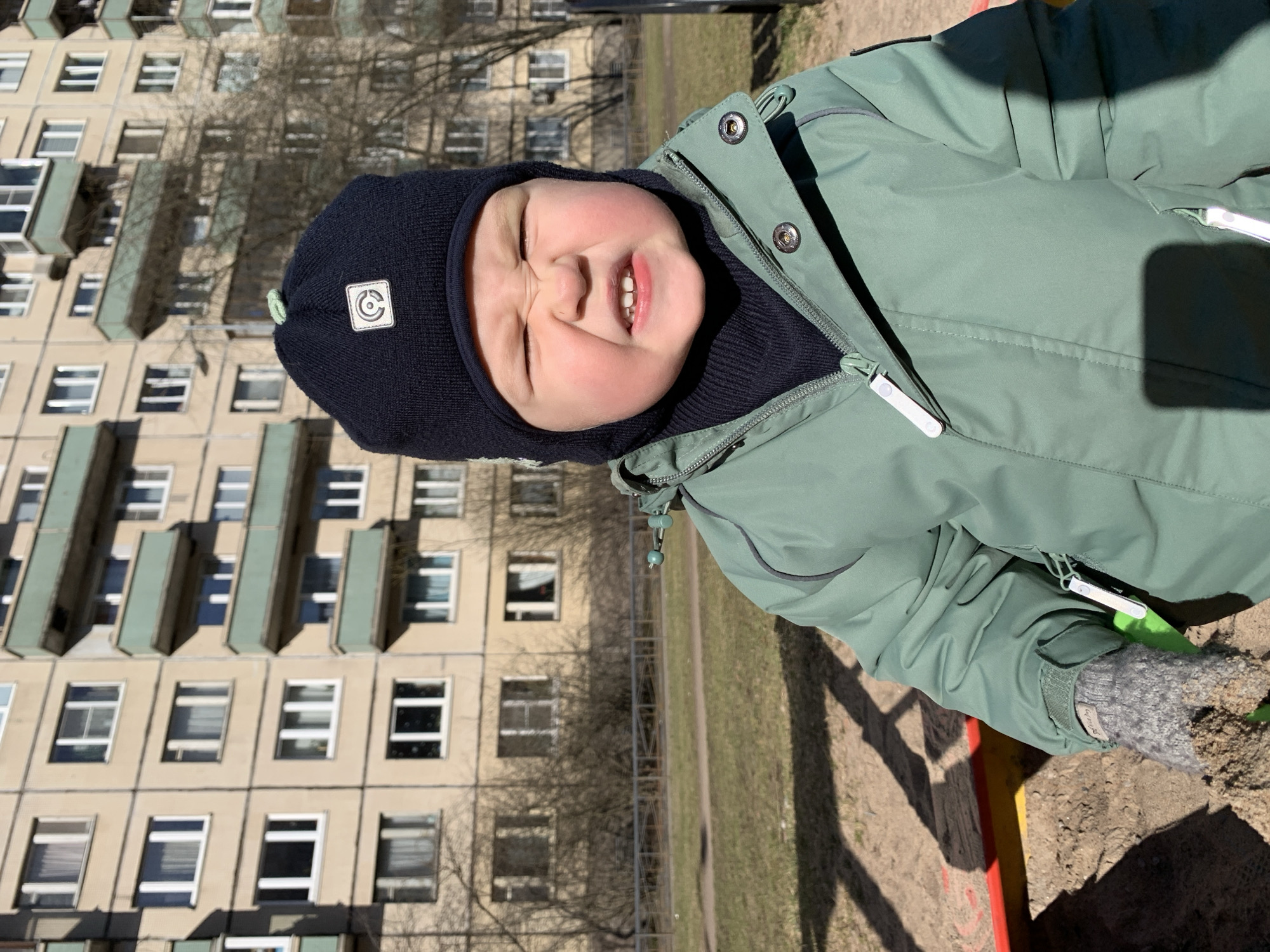 A small child in a green jacket and dark blue knit hat is making a face at the camera. The child is wearing a grey mitten. The child is outside near a sandbox, and behind the child is a multi-story building in the city of Saint Petersburg, Russia.