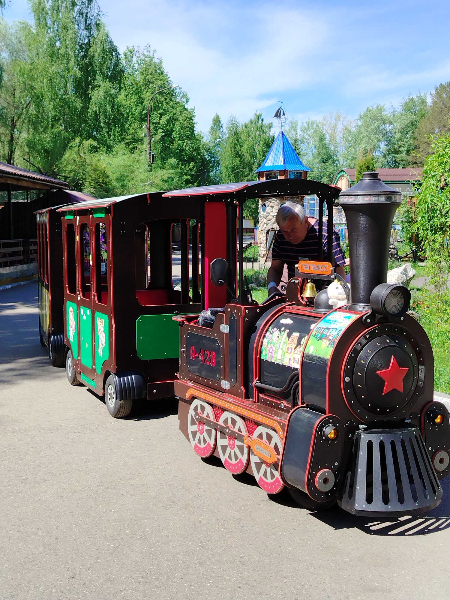 A medium-shot captures a man standing next to a miniature train at an unspecified location in Chaikovsky, Russia. The train, composed of a locomotive and several passenger cars, is predominantly brown with red accents. The front of the locomotive features a large red star. The man, middle-aged with short gray hair, is wearing a striped shirt. He appears to be inspecting or interacting with the train. The train is parked on a paved surface, likely a sidewalk or path, surrounded by greenery and trees. In the background, there are glimpses of buildings with architectural details, including a blue-roofed turret-like structure. The sky above is light blue, partially covered with light clouds. The scene suggests an amusement park or recreational area.