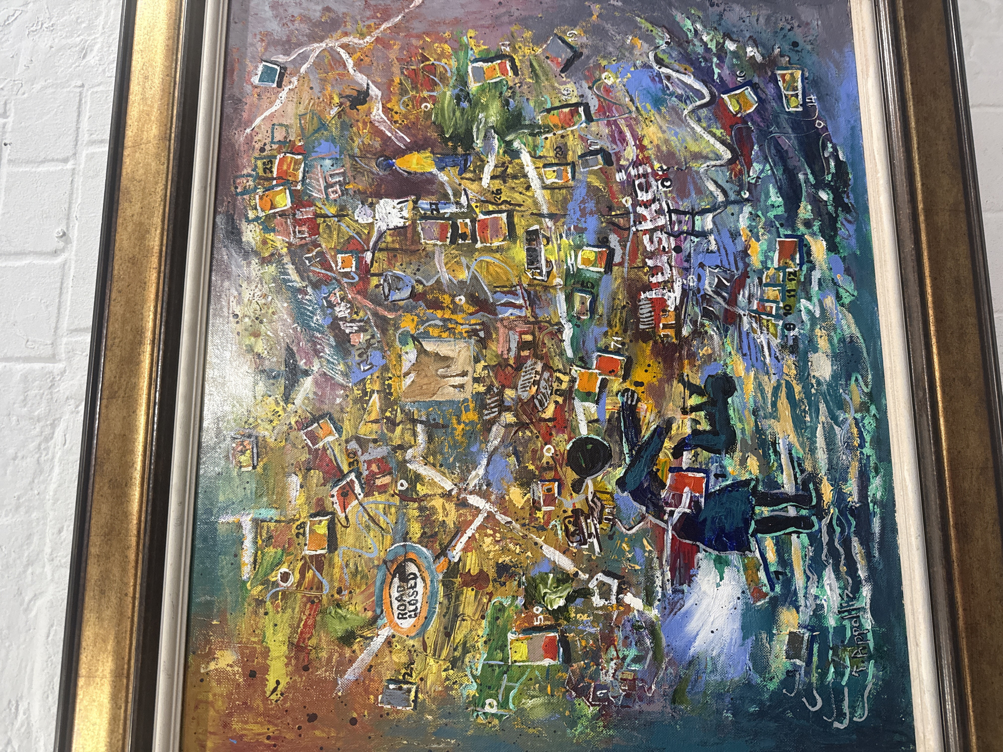 The image is of an abstract painting in a gold frame hanging on a white brick wall. The painting features a chaotic composition with a mix of shapes, lines, and colors, primarily yellows, reds, blues, and greens. There are rectangular forms scattered throughout, some resembling buildings or structures. Near the bottom left, a circular sign reads "ROAD CLOSED". A dark silhouette of what appears to be a seated figure is present in the upper right section. The signature "T. Appollis" is visible in the lower right corner of the painting. The painting's style and subject matter suggest a possible depiction of a cityscape, potentially Cape Town based on the artist's name.