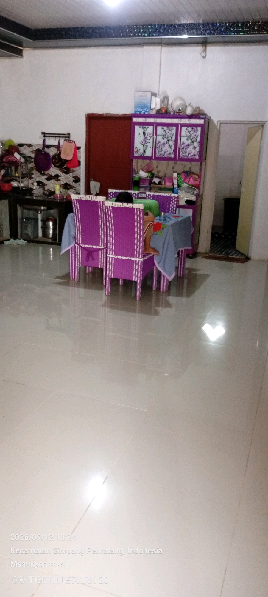 The image shows an indoor scene, likely a dining area or a multipurpose room, with a person sitting at a table. The room appears to be furnished with a purple dining set, including chairs and a table covered with a blue tablecloth. A child, possibly a toddler, is seated on one of the chairs, facing away from the camera. Behind the table, a cabinet with floral patterns is visible, along with various items on top of it. To the left, a kitchen counter with hanging utensils is present. The floor is tiled and reflective. The timestamp in the lower left corner indicates the image was taken on 2026/09/12 at 13:24 in Kecamatan Simpang Pematang, Indonesia.
