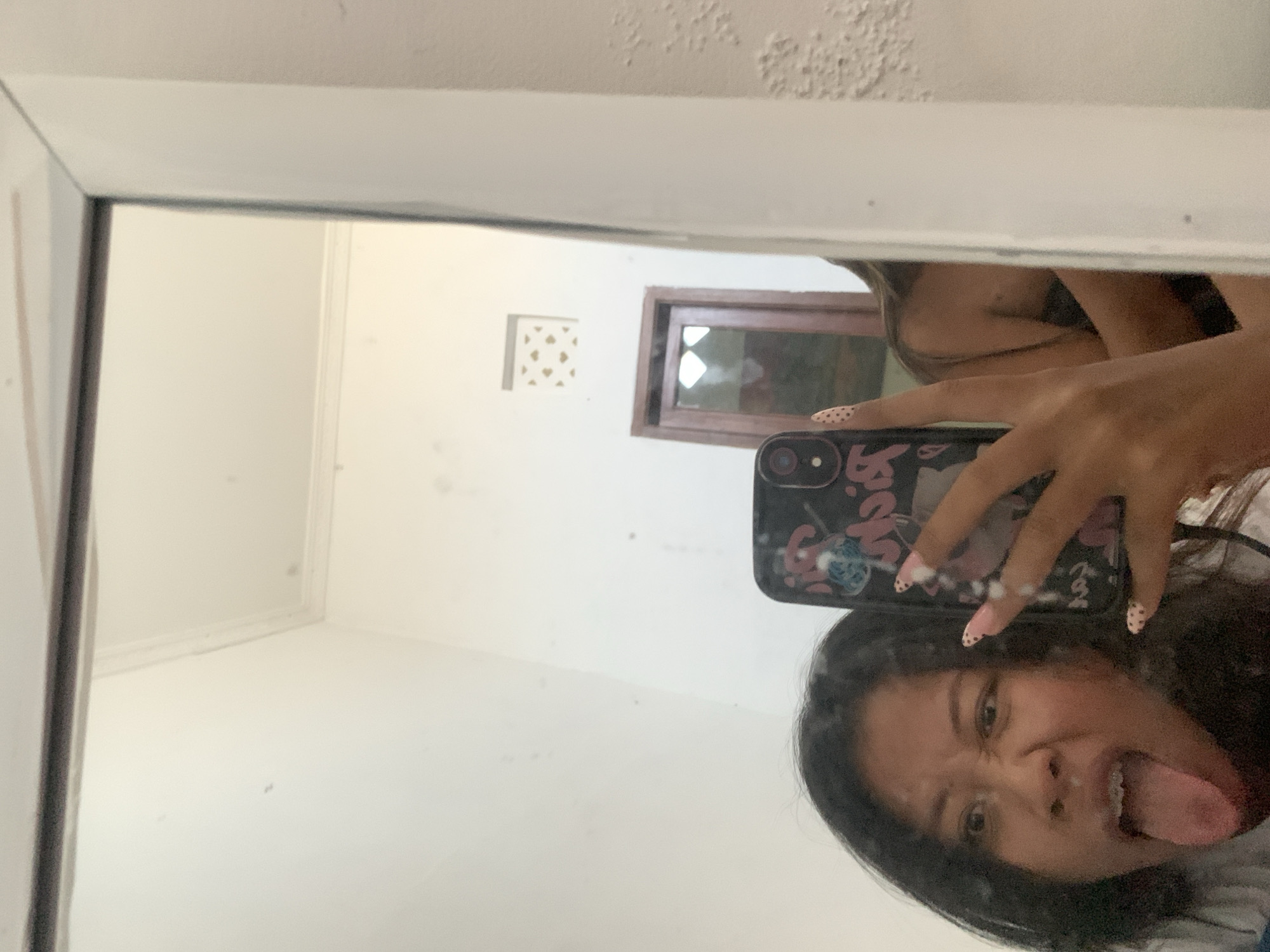 A young woman is taking a selfie in a mirror. Her face is contorted in a playful expression, with her tongue sticking out and eyes wide. She is holding a phone with a patterned case in her right hand, and her left hand is visible in the reflection. Her fingernails are painted with a light-colored polish and black polka dots. Her dark hair is loose around her shoulders. In the background, a doorway with a wooden frame is visible. The room appears to be an interior space with white walls. The lighting suggests daytime. The location is in the city of Sukawati, Indonesia.