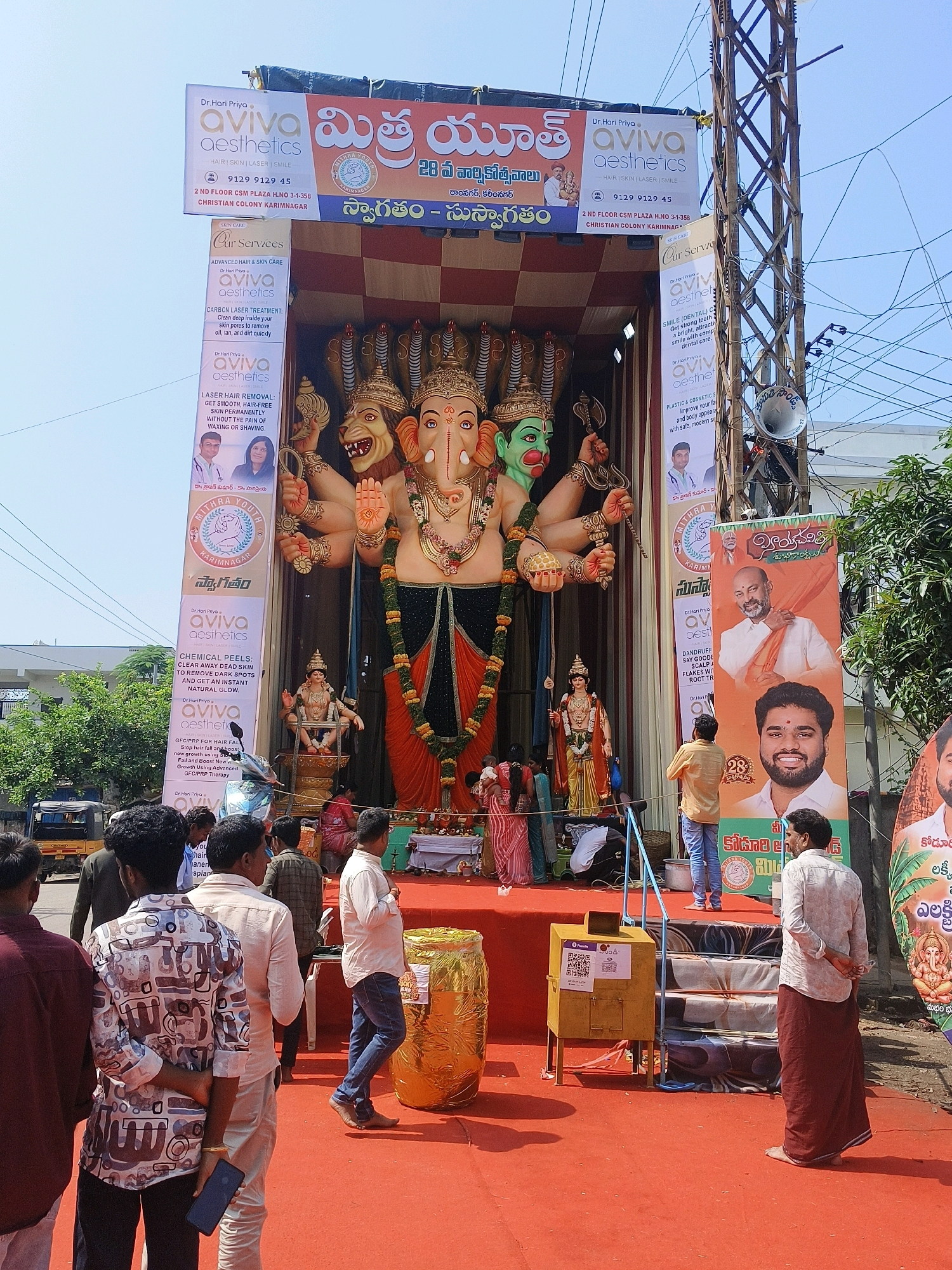 A large statue of the Hindu deity Ganesha, adorned with multiple arms and a lion's head, is prominently displayed on an elevated platform covered in red carpet. The statue is surrounded by smaller figures and decorative elements. Several banners featuring advertisements and political figures are hung on either side of the statue. In the foreground, a group of men are gathered, some observing the statue and others interacting with their surroundings. A yellow donation box with QR codes is visible near a set of stairs. The scene appears to be an outdoor public event, likely a religious festival or celebration, taking place in the city of Karimnagar, India, as indicated by text on the banners. The environment suggests a sunny day, with a clear sky and urban infrastructure visible in the background, including power lines and buildings.