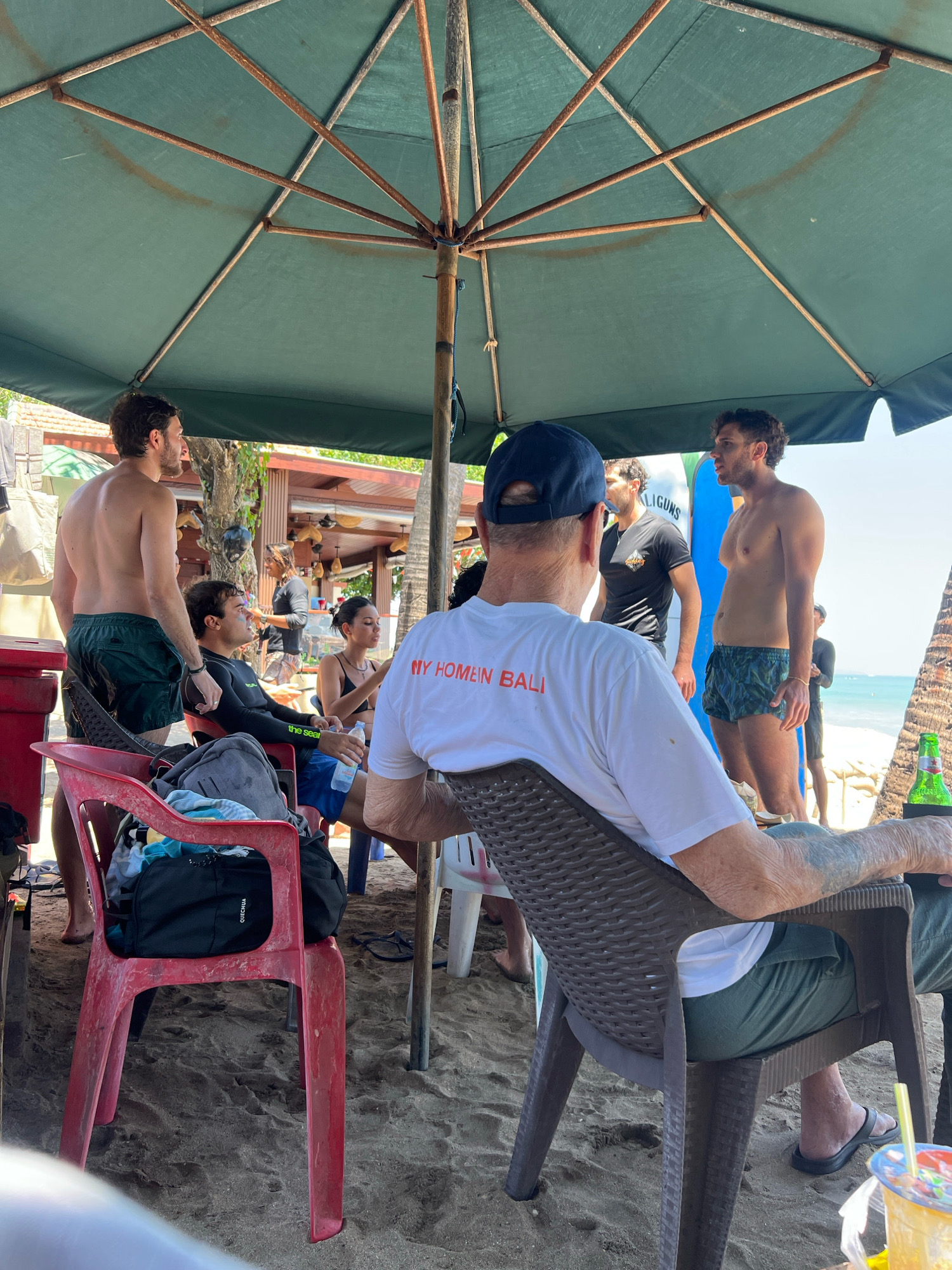 A group of individuals are gathered under a green umbrella on a sandy beach in Kuta, Indonesia. In the foreground, a person wearing a white t-shirt with "MY HOMEYN BALI" printed on the back is seated in a brown chair, facing away from the camera. To their left, a red plastic chair holds a black backpack. To the left of that, a shirtless man with dark hair and green shorts stands. In the middle ground, a man in a black wetsuit is seated on a red chair, holding a bottle. To his right, a woman in a black bikini is seated and holding a water bottle. Further back, a man in a black t-shirt is standing, and another man in a dark wetsuit is standing behind him, looking out towards the ocean. The background features the ocean and palm trees.
