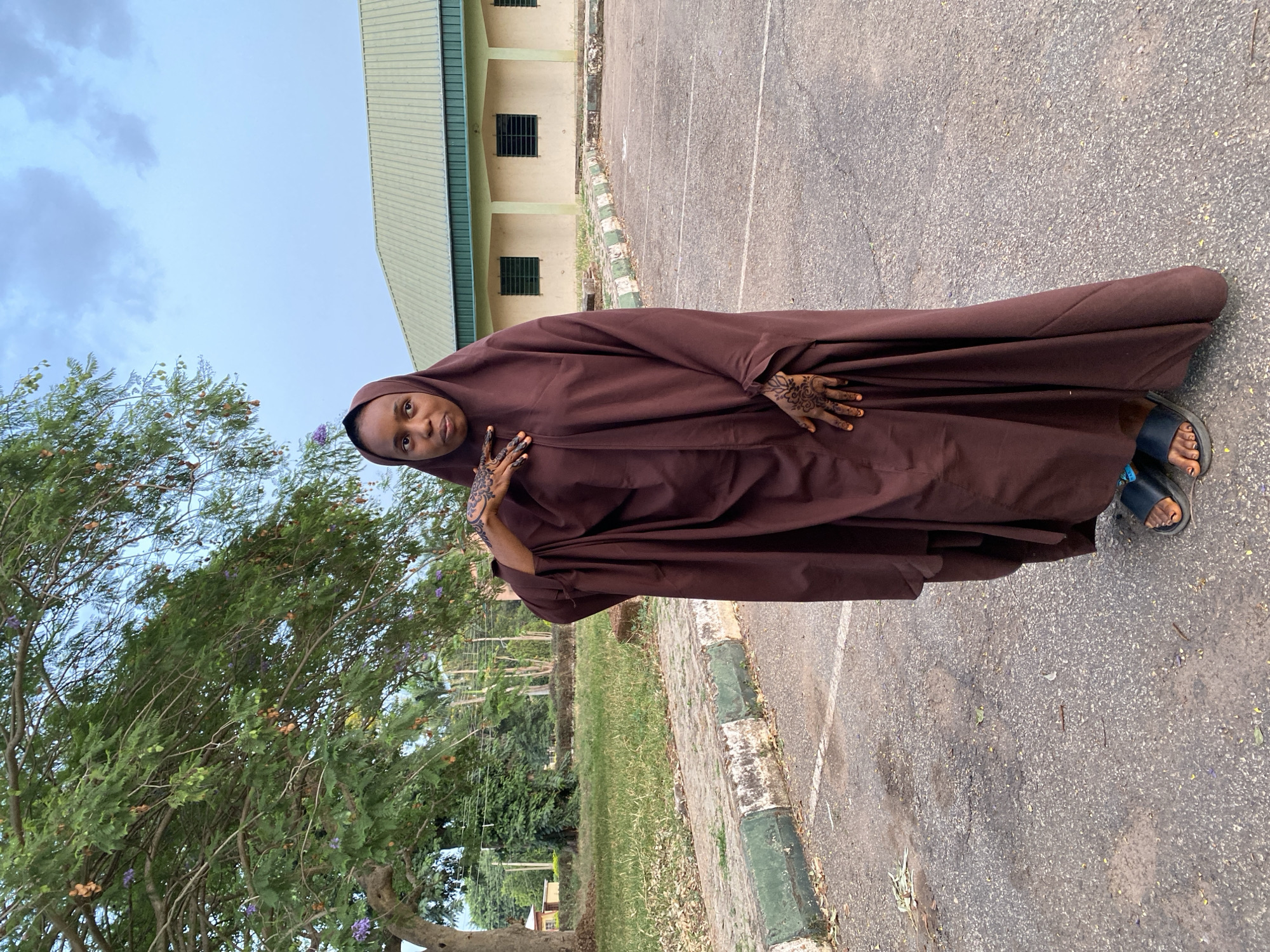 A woman is standing outside on a paved path in Vom, Nigeria. She is wearing a brown hijab with henna on her hands and blue sandals. To her left is a tree. Behind her is a grassy lawn with some trees and bushes, and then a low building with a green metal roof. The sky is overcast.
