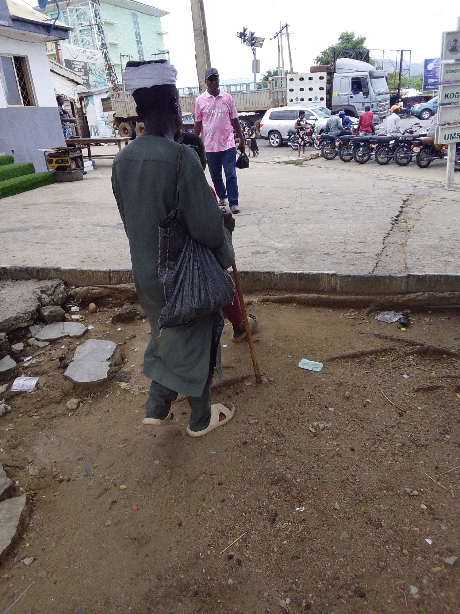 This image captures a bustling street scene in Lokoja, Nigeria, under what appears to be daytime conditions with ambient lighting.
In the foreground, a man is seen from behind, wearing a dark green traditional tunic or robe, dark trousers, and light-colored sandals. He has a white head covering and carries a dark, possibly woven or plastic, bag slung over his left shoulder. He holds a long wooden staff or cane, suggesting support for walking or a traditional accessory. The ground immediately beneath him is unpaved, dusty, and uneven, littered with small stones, debris, and broken pieces of concrete, exposing the earth and roots underneath.
The midground reveals a paved road or sidewalk area. To the right of the foreground man, another man walks away from the camera, wearing a pink polo shirt with "Emirates FLY BETTER" visible, blue jeans, and a dark baseball cap. Behind them, traffic and people are visible. A white SUV is stopped or parked, and a line of motorcycles (okadas) are parked alongside the road, with several riders seated on them. A large grey truck, possibly a flatbed or semi-truck, is prominent, carrying large cages or crates. Utility poles with wires and traffic lights are also present.
On the left side of the midground, a white building with "SUIT RIA HOTELS" is visible, along with other commercial advertisements. A yellow portable generator is placed near the building's entrance. The background shows more buildings, some with a light green or blue hue, suggesting an urban or commercial environment. Distant hills or mountains are faintly visible on the horizon. Partially obscured street signs on the right side include text like "KOGI" and "UMS."
The scene depicts a typical urban environment with a mix of pedestrian and vehicular activity, varied infrastructure (paved roads alongside unpaved, damaged sections), and commercial establishments. The overall impression is one of a busy, functional street.