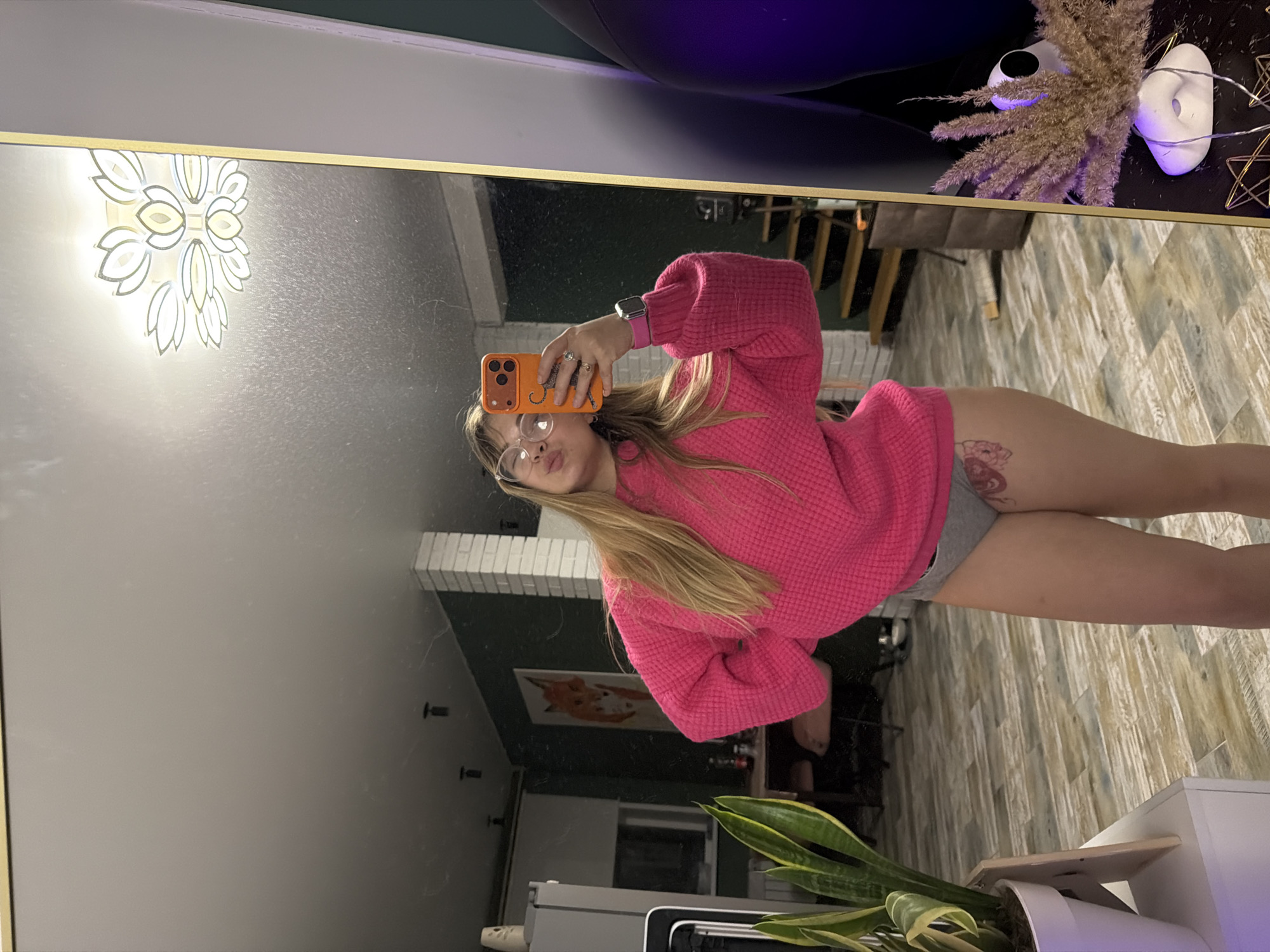 The image shows a mirror selfie of a woman in Большая Шильна, Russia. She is wearing a pink sweater and gray shorts. Her hair is blonde, and she wears glasses. She is holding an orange phone. The background includes a white brick wall, a picture of a fox, a plant in a white pot, and a wood-paneled wall.