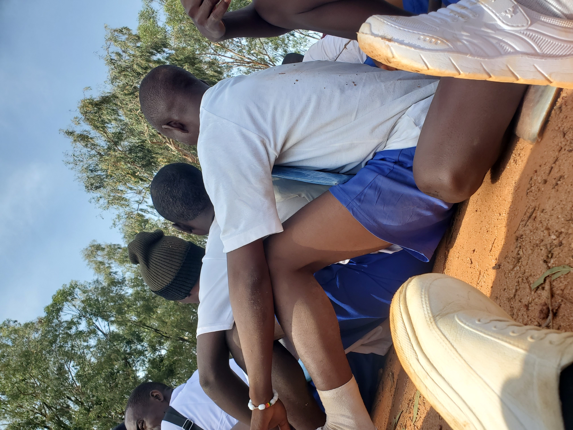 This media file depicts a group of young men, likely students, sitting outdoors on a sunny day. The scene is set in a rural or semi-rural location in Barakin Rafin Gora, Nigeria, with a backdrop of trees and a clear blue sky. The individuals are wearing casual attire, including white t-shirts and blue shorts, with one person wearing a dark beanie.
The subjects appear to be engaged in a moment of rest or anticipation, possibly before or after an activity. Their postures suggest a relaxed atmosphere. Notable details include the worn white sneakers visible in the foreground, indicating the practicality of their footwear. The ground appears to be dirt, further suggesting an outdoor, possibly recreational or educational setting. The lighting suggests it is daytime, likely mid-morning or afternoon. The overall mood appears casual and communal. There is no visible text in the image.