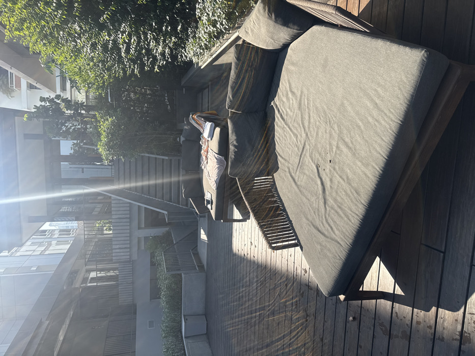 The image displays an outdoor leisure area on a bright, sunny day, consistent with a modern hotel or residential complex in Bangkok, Thailand. The foreground contains a large, empty, roundish outdoor daybed featuring dark grey cushions and a woven frame. To its right, a rectangular outdoor chaise lounge, also with dark grey cushions, is occupied by a light pink garment or towel, a folded white towel, and a dark mug placed on a small coaster. The ground consists of a wooden deck, exhibiting distinct sunlit and shadowed sections. In the mid-ground, a prominent concrete staircase with metal railings ascends towards a building facade featuring large glass doors. Another set of stairs descends to a lower level on the left. Dense green foliage, comprising bushes and small trees, frames the right side of the scene and extends behind the architectural elements. Modern multi-story buildings, characterized by concrete and glass construction, are visible in the background. Strong sunlight creates visible lens flare in the upper-center portion of the image. No individuals are present in the scene.