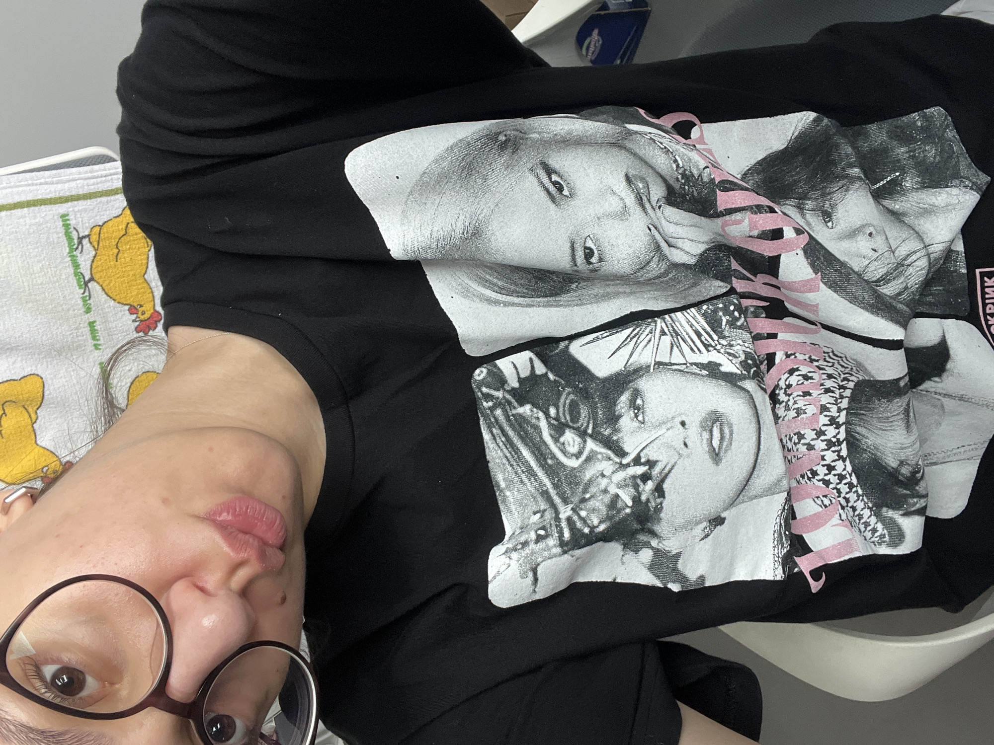 This is a close-up, indoor selfie of a young woman wearing a black t-shirt with a graphic print. The print features black and white portraits of what appear to be members of the K-pop group BLACKPINK, with the text "BLACKPINK GIRLS" in pink. The woman has on glasses and is making a pouty face. A yellow towel with a drawing of a chicken is visible in the background. The image does not provide enough information to determine a specific location within Ruse, Bulgaria.