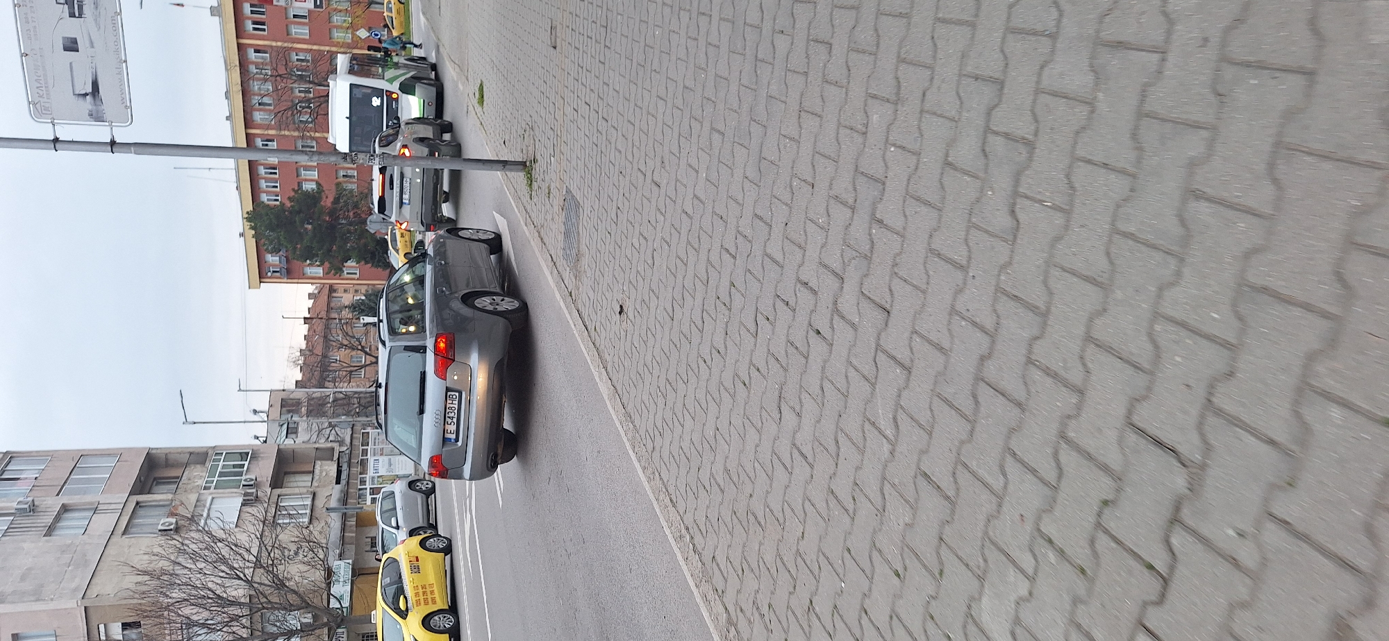 The image depicts a street scene in Blagoevgrad, Bulgaria. The road is lined with parked cars, including a grey Audi with license plate "E 5438 HB" and a yellow taxi. The buildings in the background are residential apartments. A sidewalk paved with gray bricks borders the road. A light pole and street sign are visible on the left side of the image. The sky is overcast.