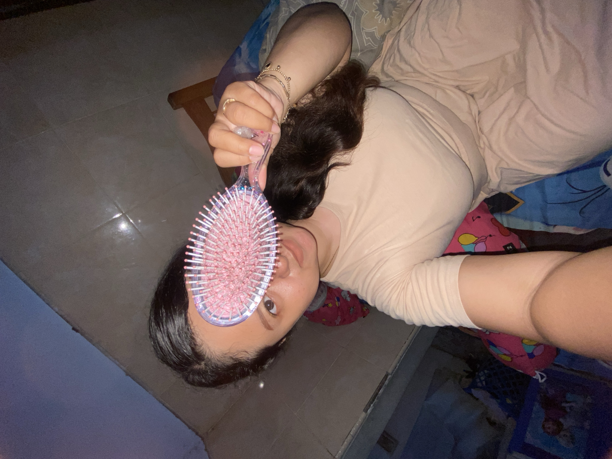 A person is lying on a bed in an interior space. The person, possibly female, is wearing a beige colored outfit and has brown hair. The person is holding a pink hairbrush in front of their face. The background includes a blue and pink patterned bedsheet and a dark tiled floor. The location is not specified.