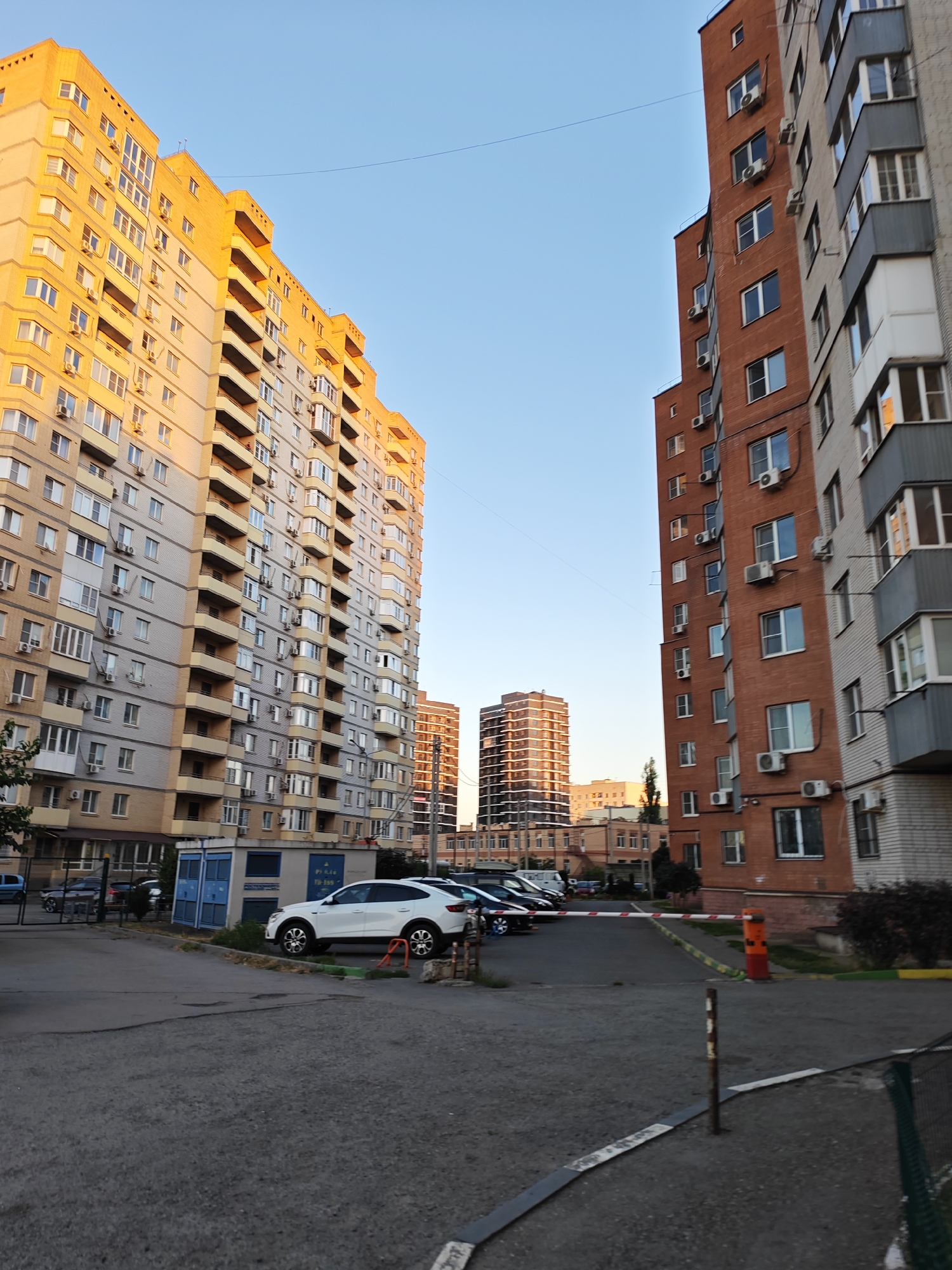 In Bataysk, Russia, the scene captures an outdoor urban environment at dusk. The dominant features are tall residential buildings, one in the foreground and to the left, a pale yellow and gray brick multi-story apartment complex, bathed in the warm glow of the setting sun. To the right and further back, another brick apartment building with a reddish-brown facade stands. In the mid-ground, two modern, glass-fronted skyscrapers are visible against a clear blue sky. A black SUV is parked on the asphalt. A white crossover vehicle is also parked. In the lower portion of the image, a paved area appears to be a parking lot or driveway. A red and white striped barrier is in the middle of the scene, with a wooden pole to its left. A dark green wire fence runs along the right edge of the image. The overall impression is one of a residential urban landscape at the end of the day.