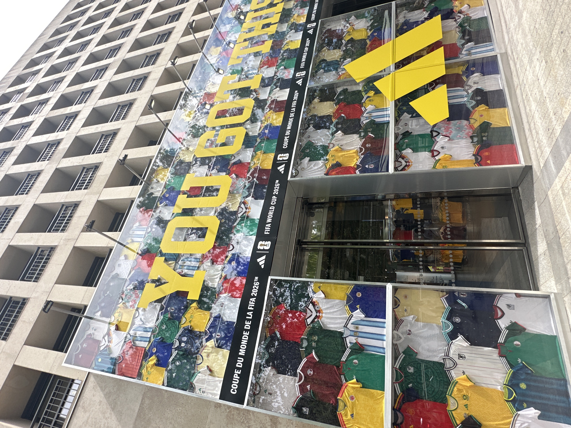This image depicts the exterior of an Adidas store in Paris, France. The facade of the building is covered in a large display of football jerseys, presumably from various national teams and past FIFA World Cup editions. Prominently featured on the upper part of the display are large yellow letters spelling out "YOU GOT THIS". Below this, text reads "COUPE DU MONDE DE LA FIFA 2026™" alongside Adidas and FIFA World Cup logos. The store entrance, with glass doors, is visible in the center. The building itself is a multi-story structure with a grid-like facade of windows and balconies. The scene is captured from a street-level perspective on a bright, daytime.