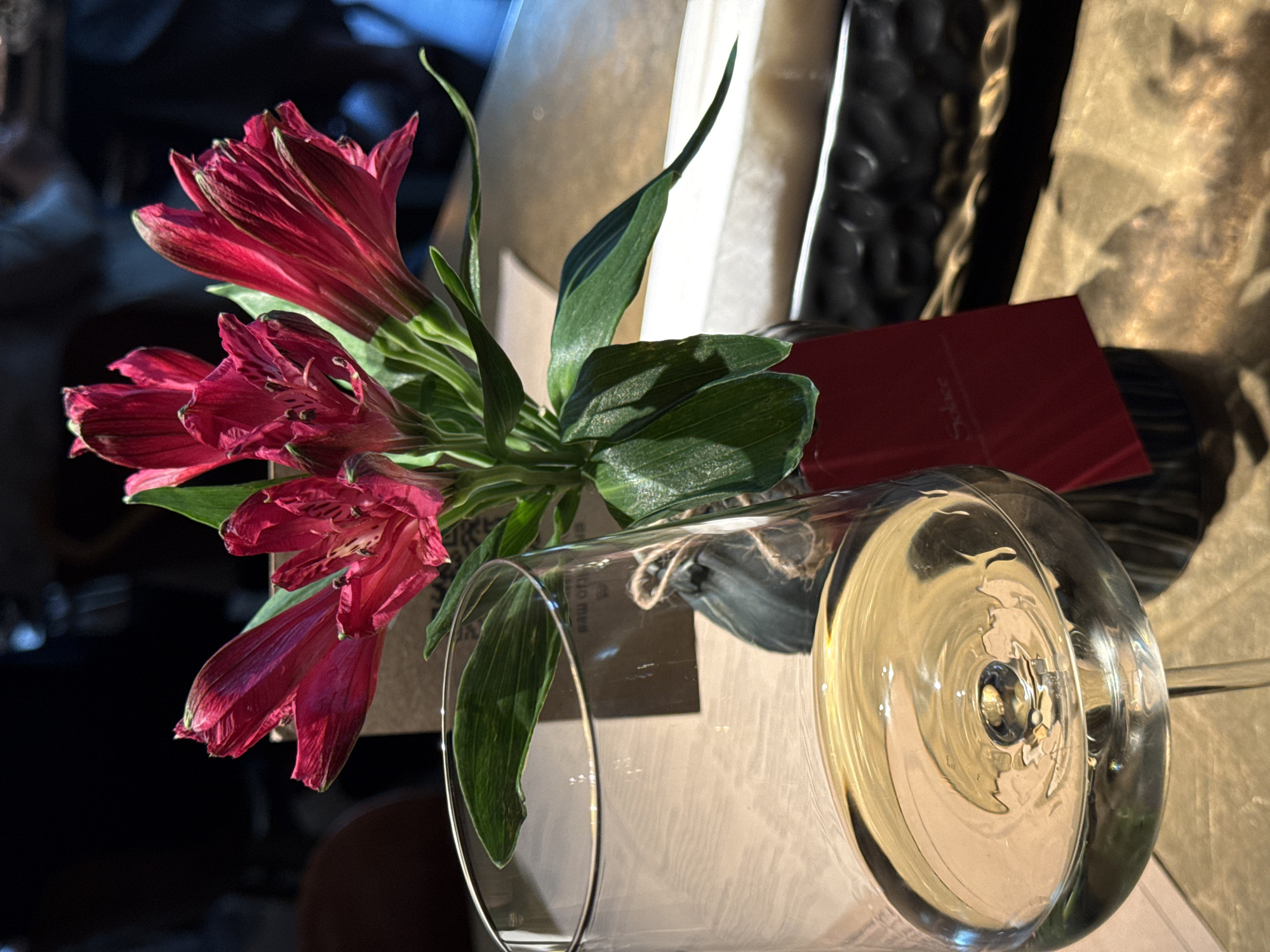 A dimly lit still life featuring a bouquet of red Alstroemeria flowers in a clear glass vase. A small red card is situated near the vase, partially obscured. The location is Kazan, Russia, suggested by the "Sultse" label on the card. The background includes textured materials and a glossy white surface. A portion of a person is visible in the far background to the left. The vase is mostly full of a light colored liquid. The arrangement sits on a surface with a light, patterned texture.
